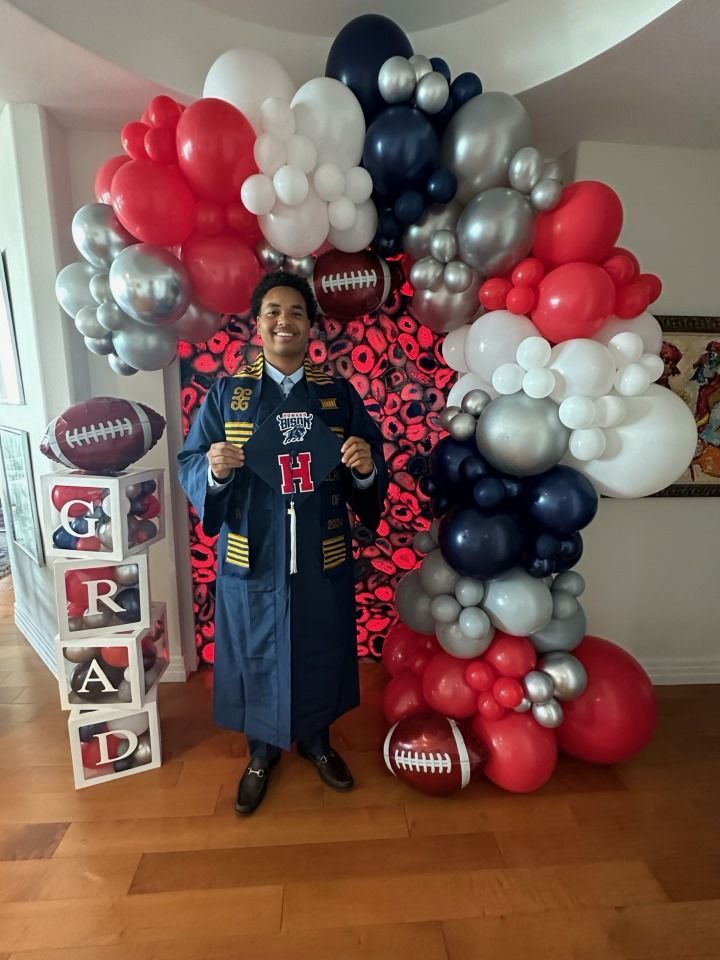 A man in a graduation cap and gown is standing in front of a balloon arch.