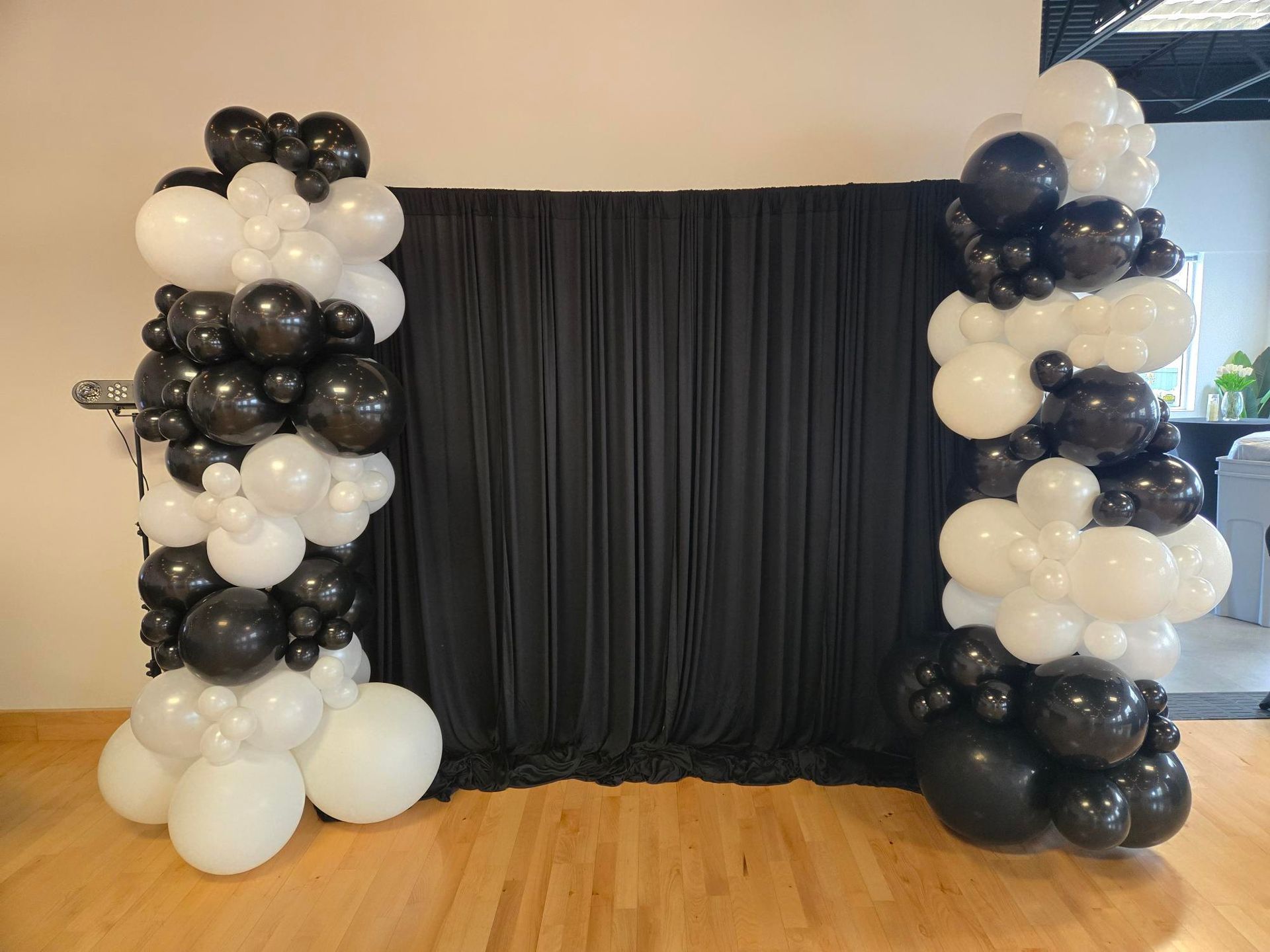 Black and white balloon columns frame a black fabric backdrop, set against a wooden floor in a room.