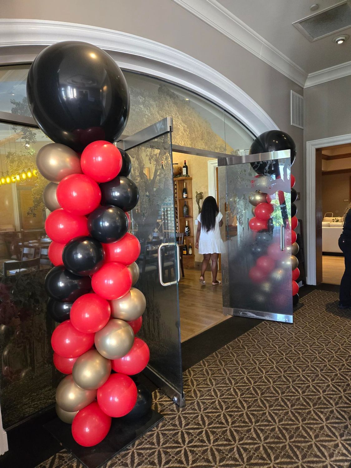 Balloon columns in red, gold, and black flank a doorway leading into a restaurant. A person in a white dress is visible inside.