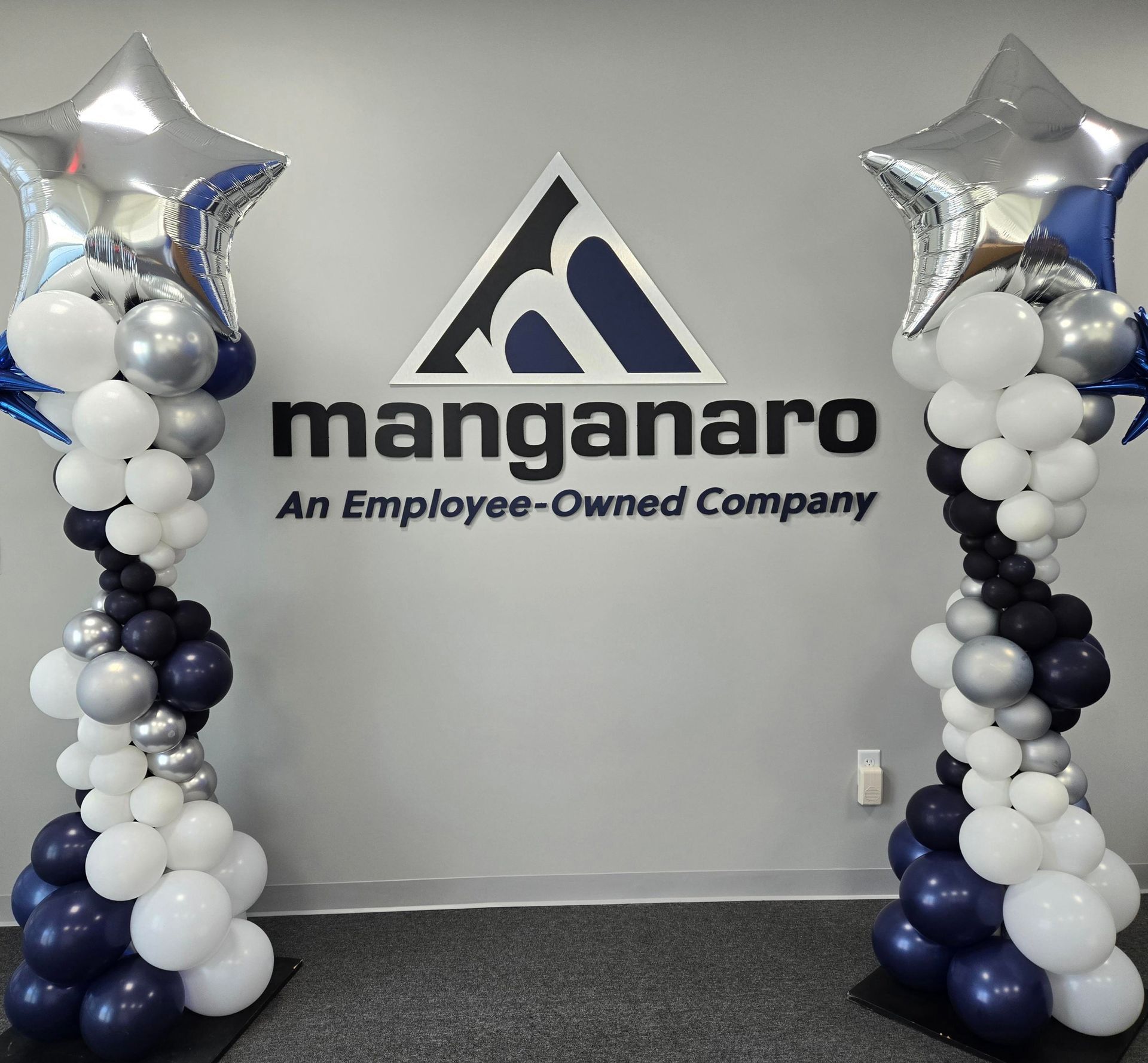 Two balloon columns flank a company logo. The columns are white, silver, and navy blue, topped with silver star balloons.