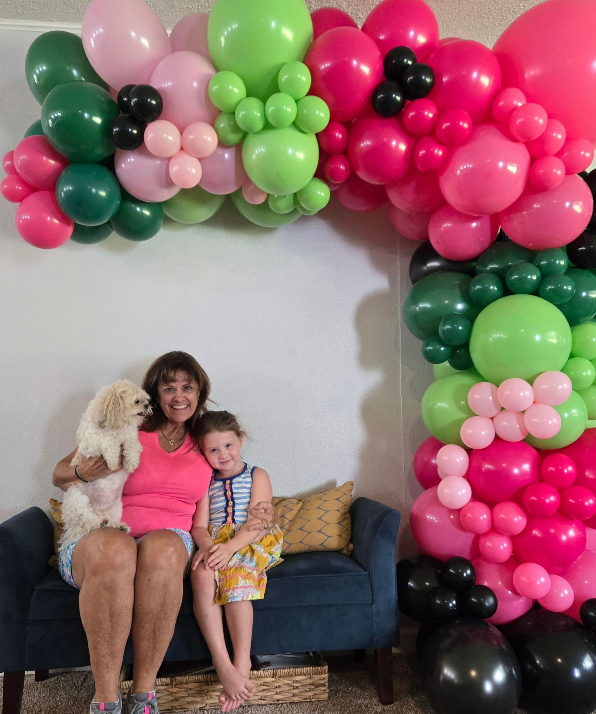 Woman, girl, and dog sit under a colorful balloon arch; pink, green, and black balloons.