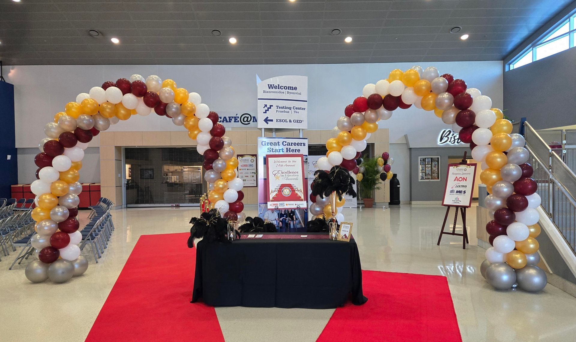 Balloons arch over a red carpet, a table with decorations, and signs inside a building.