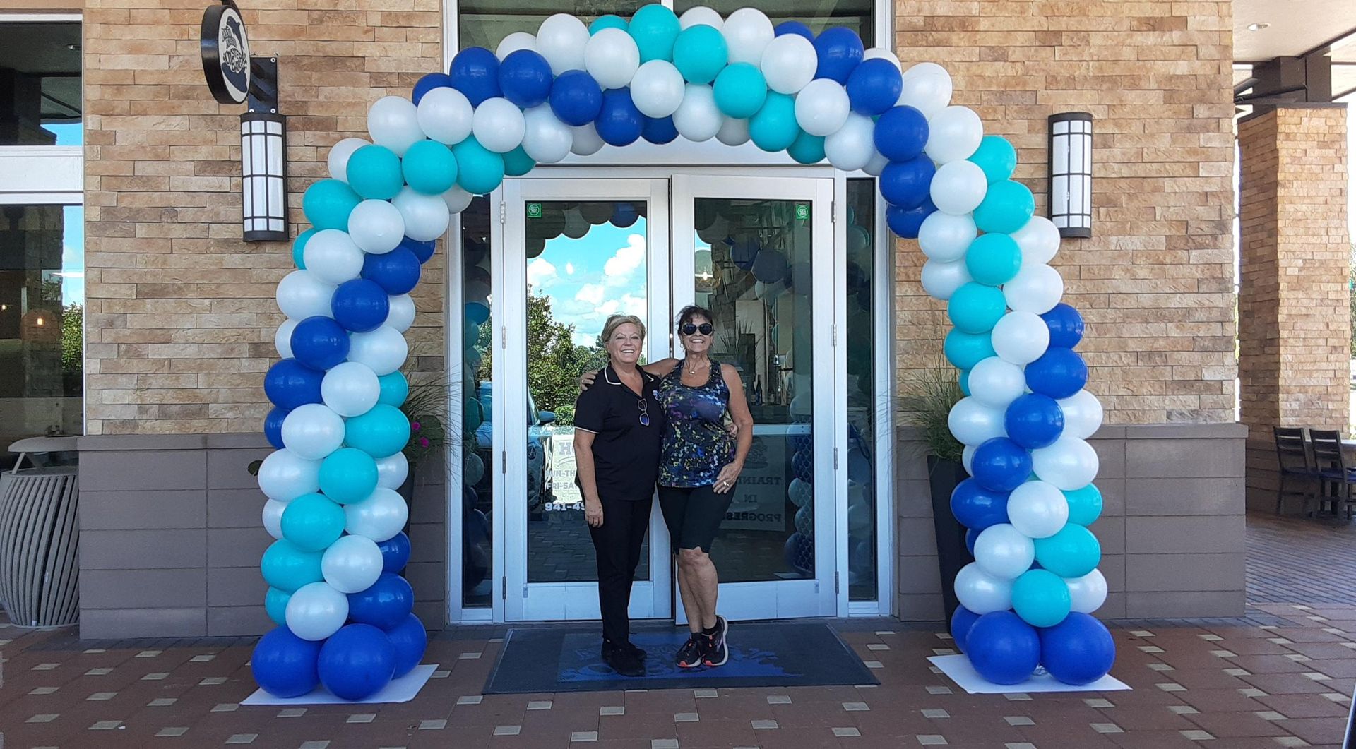Two people stand together under a decorative blue and white balloon archway at the entrance of a stone-clad building.