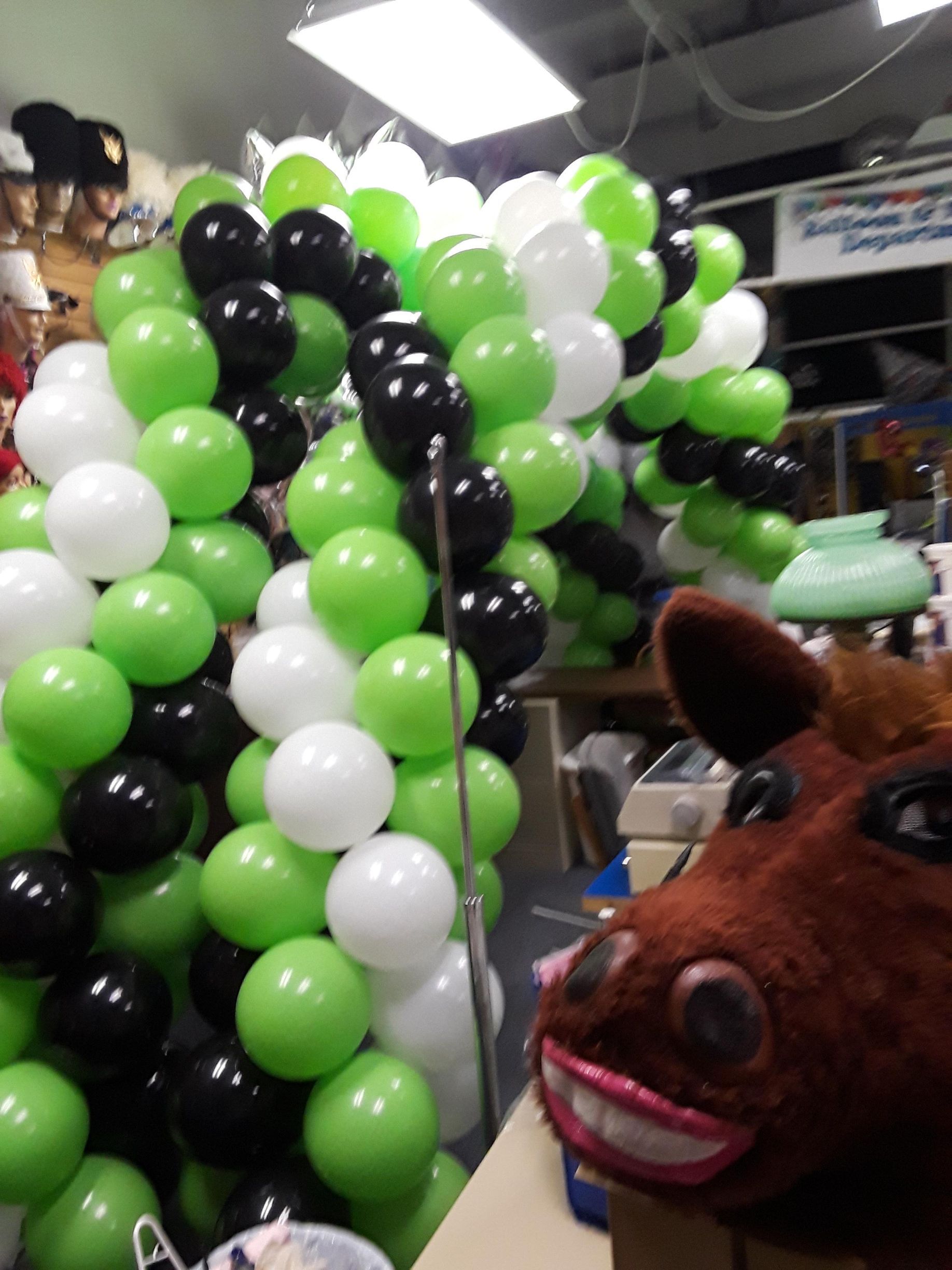 A balloon arch in green, white, and black stands behind a brown plush horse head with a wide, smiling mouth.