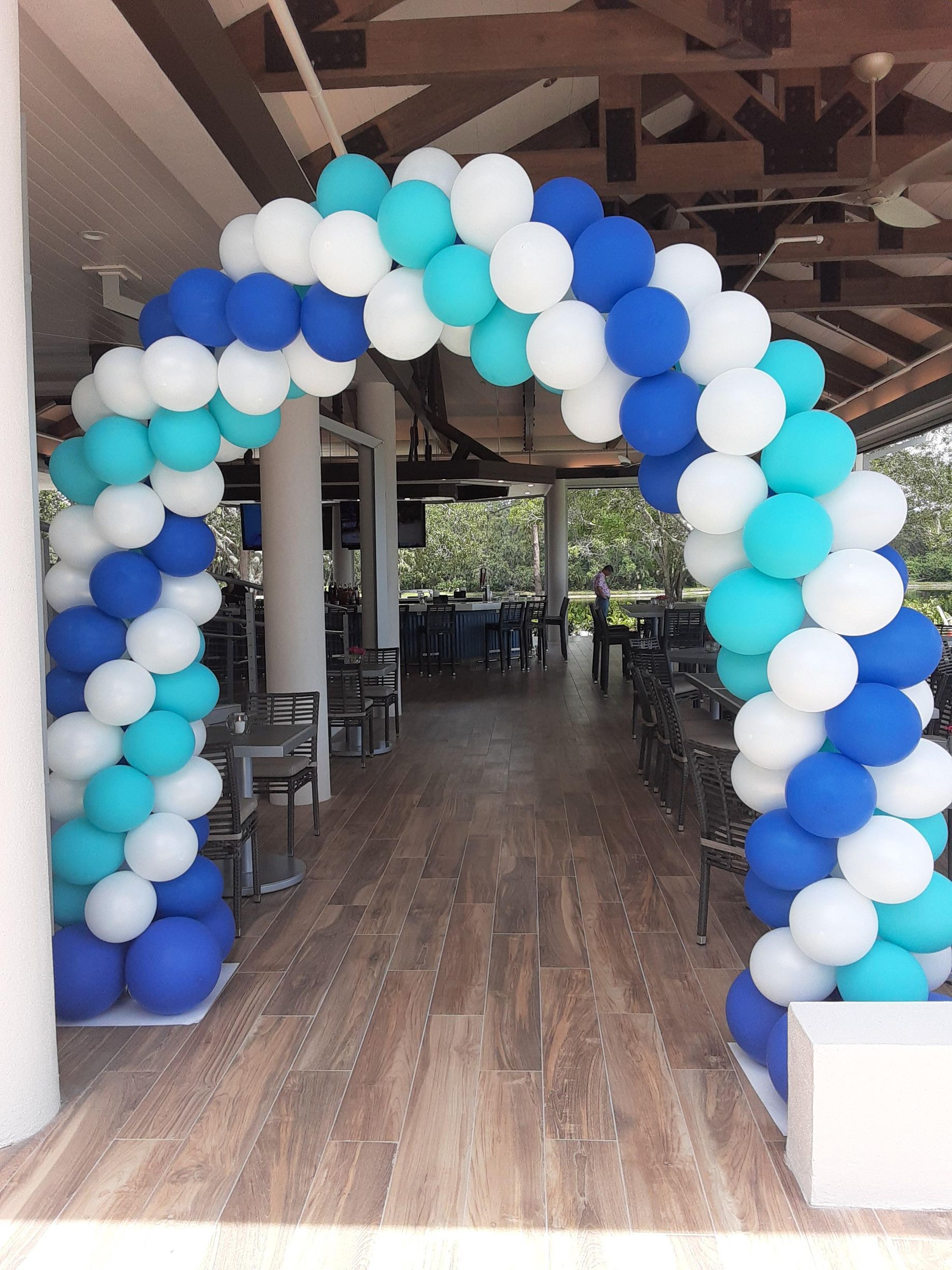A balloon arch in shades of blue and white frames a walkway entrance to an outdoor patio area with tables and chairs.