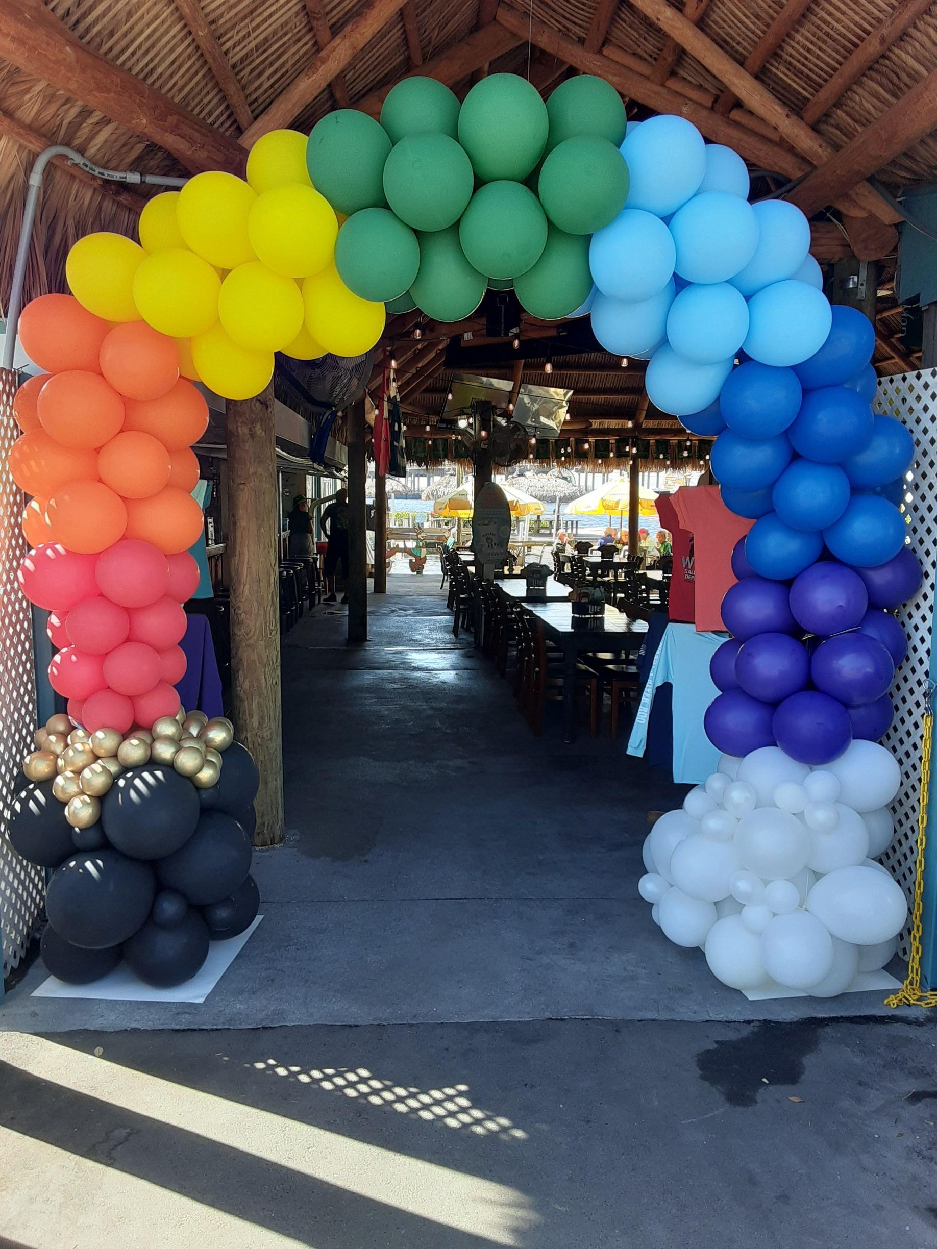 A rainbow-colored balloon arch frames an entryway, starting with black and white balloons at the base and blending colors.