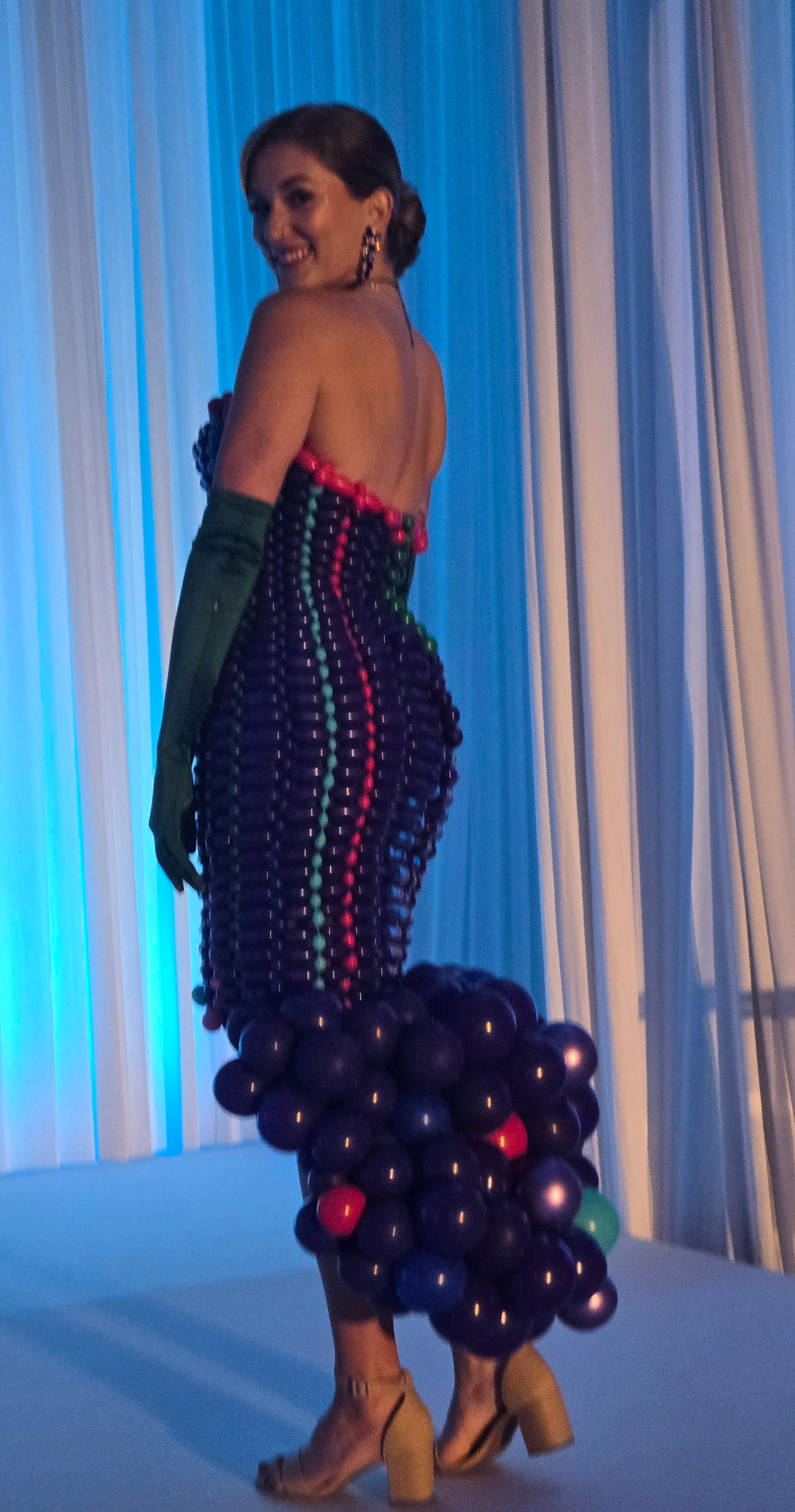 A person wears a strapless dress made of dark balloons with pink and blue accents, accessorized with long green gloves. 