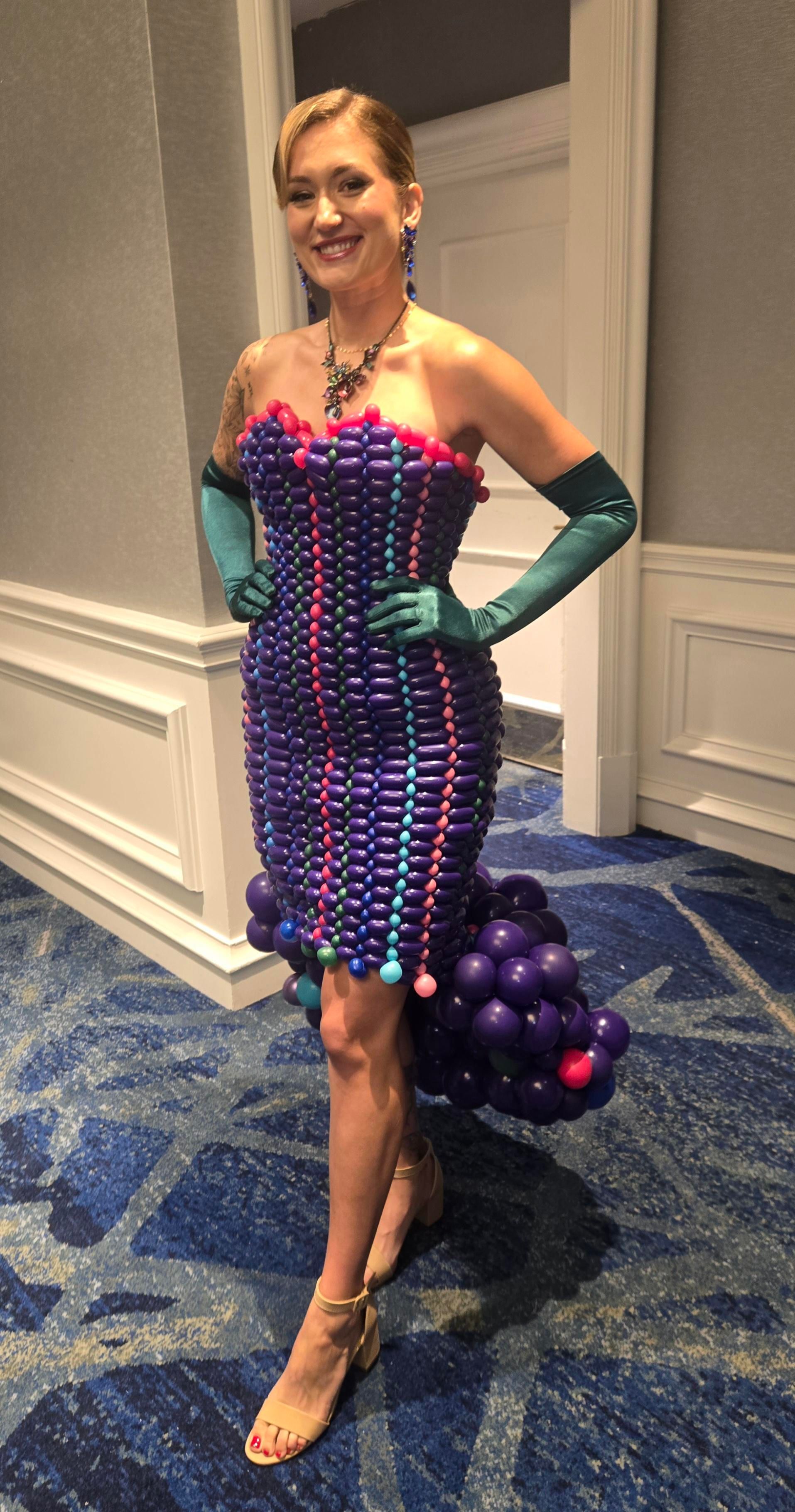 A person wearing a strapless dress made of small, purple and blue balloons with a balloon train, standing on patterned carpet. 