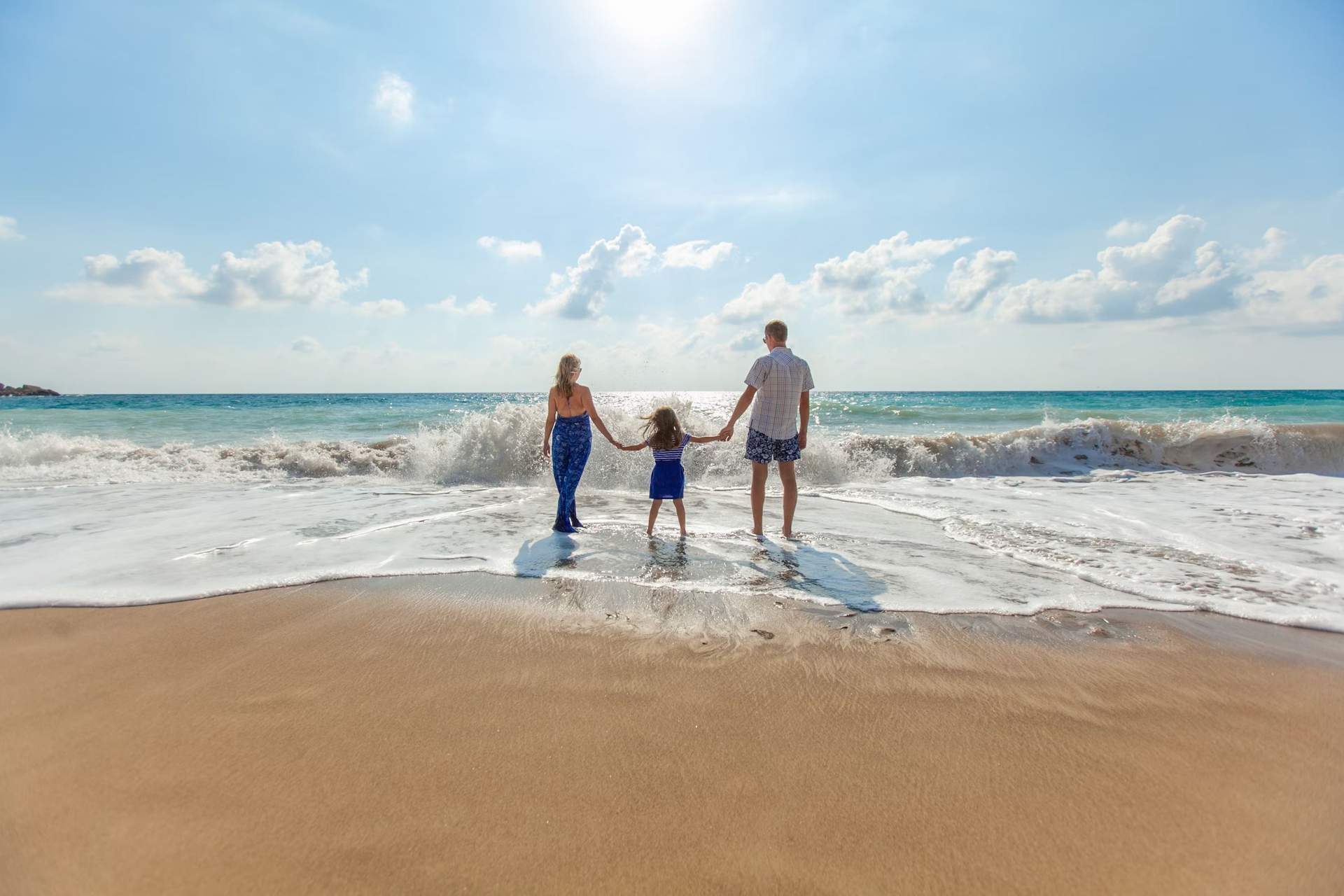 A mother, a father, and a toddler hold hands while walking along the beach at the water's edge. 