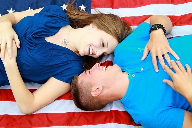 A heterosexual couple rests on an American flag blanket and smiles at each other. 