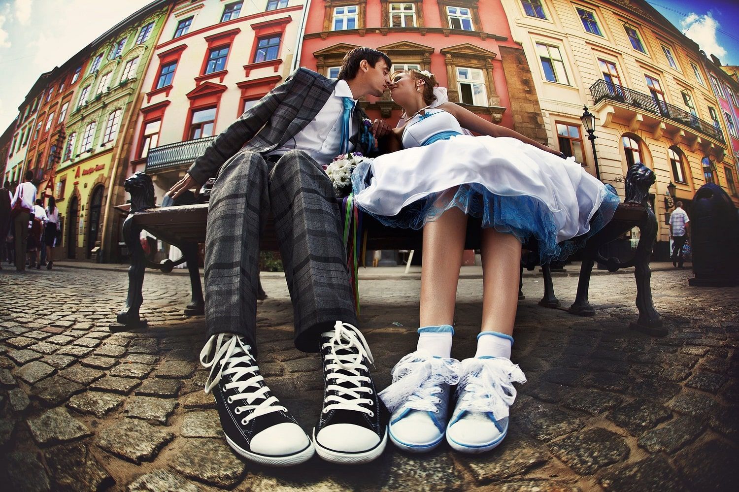 A couple wearing sneakers and formal clothing sit on a bench outside and kiss. 