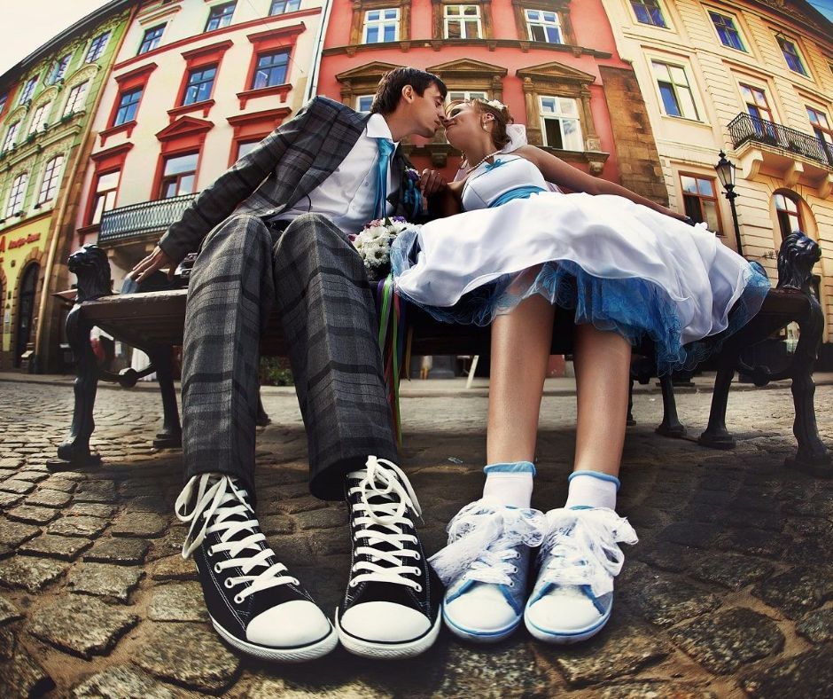 A couple wearing sneakers and formal clothing sit on a bench outside and kiss. 