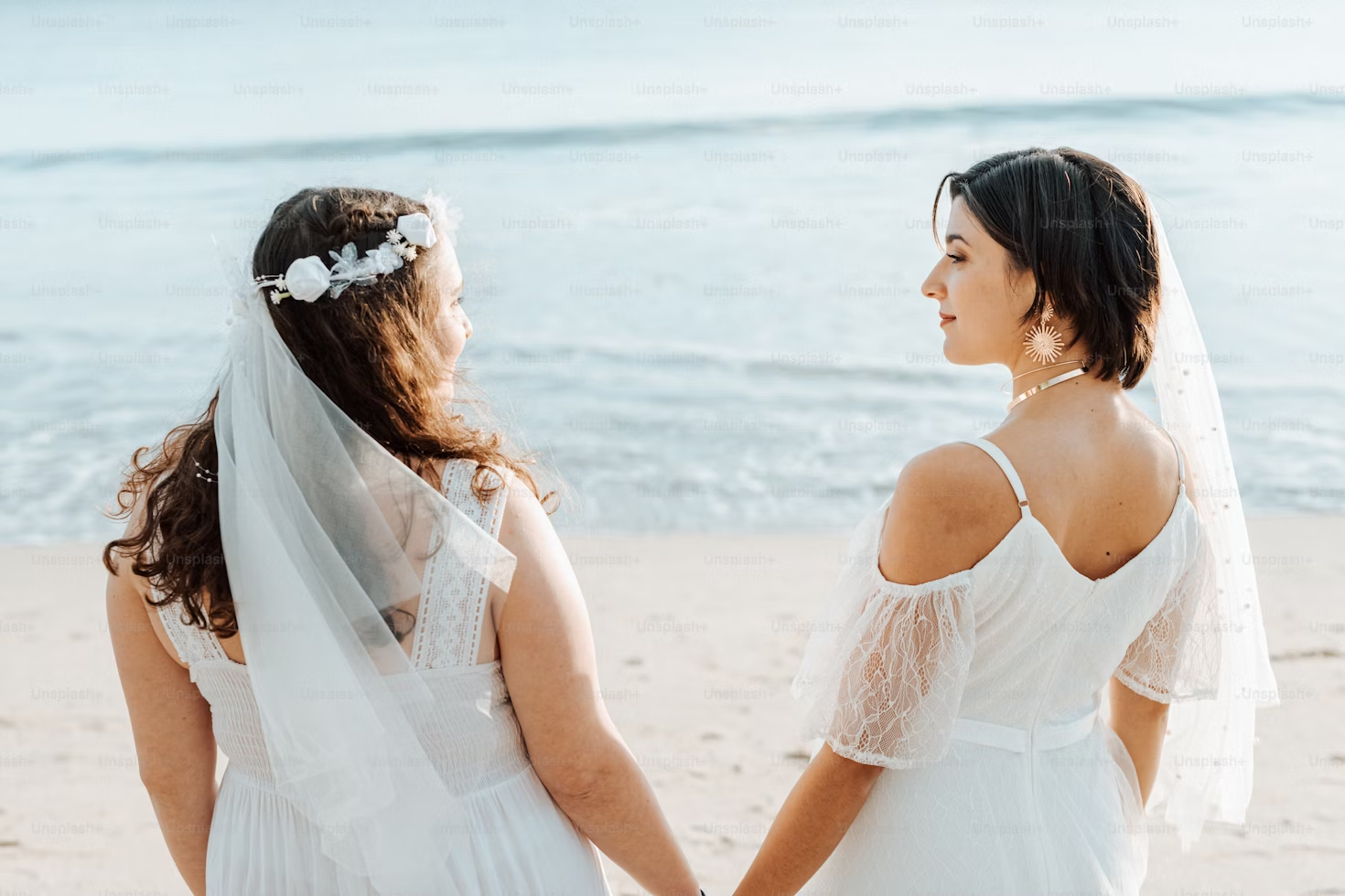 Two lesbian brides hold hands while standing on the beach in wedding gowns. 