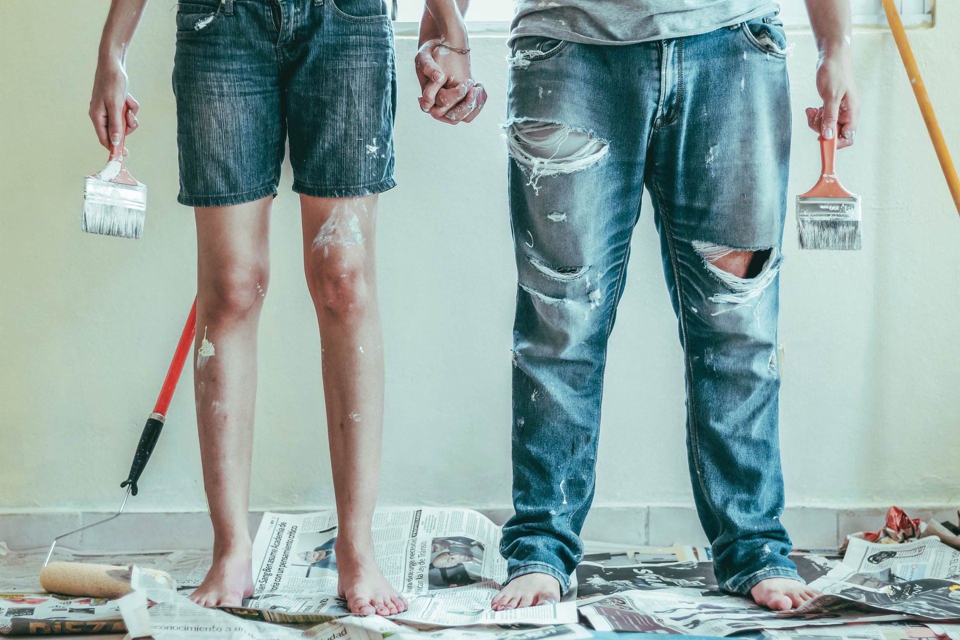 A heterosexual couple holds hands as they also hold paintbrushes to paint a room.