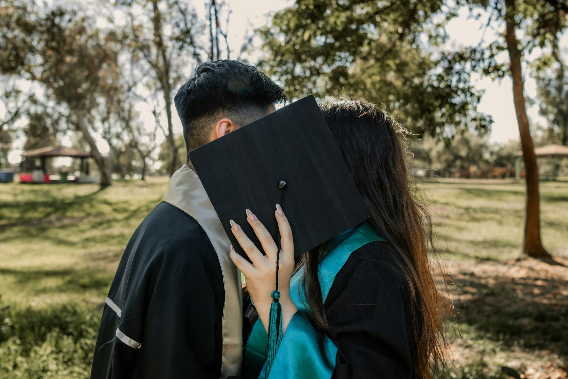 A couple kisses behind a graduation cap held in the woman's hand. 