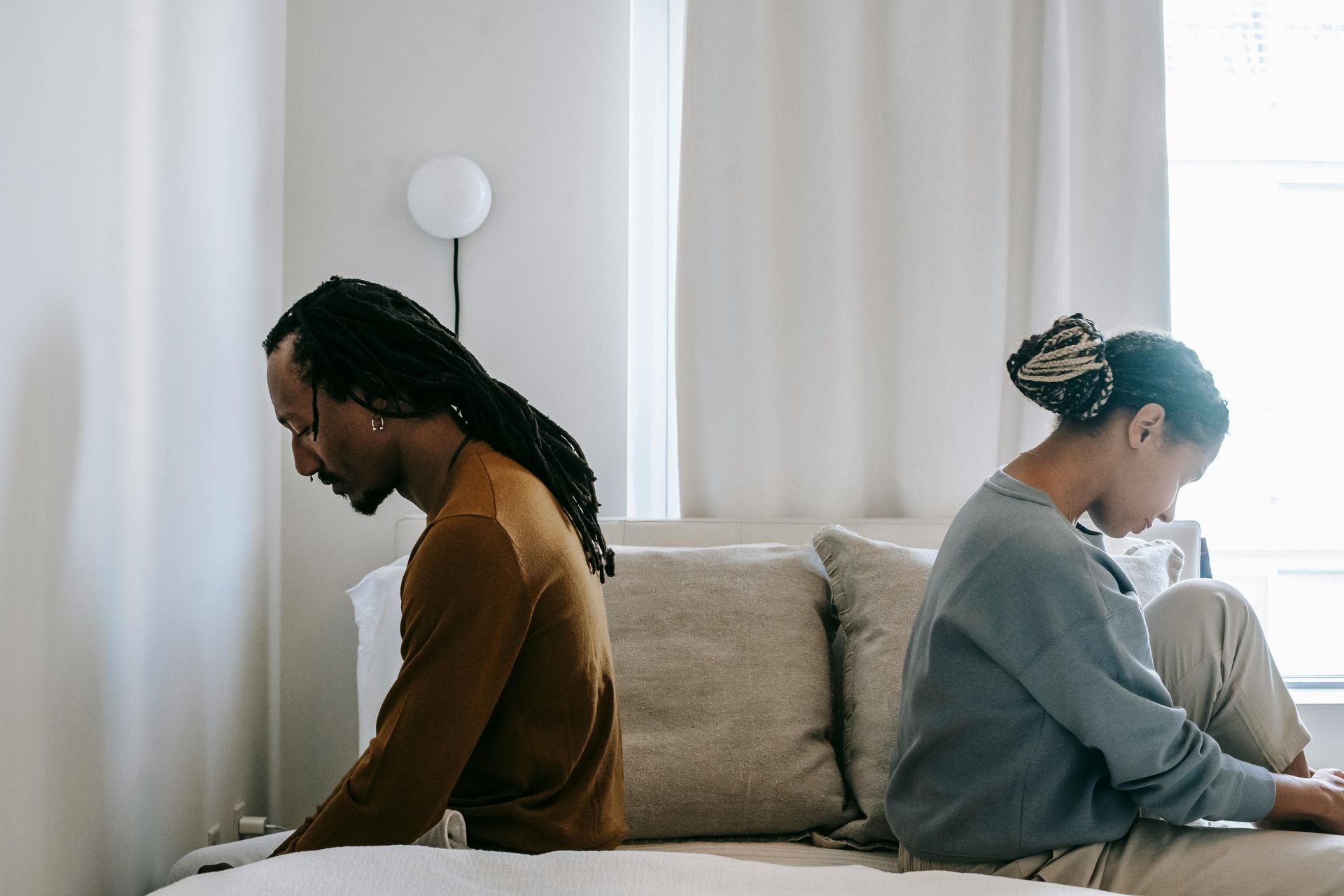 A man and a woman face away from each on a bed and look distressed.