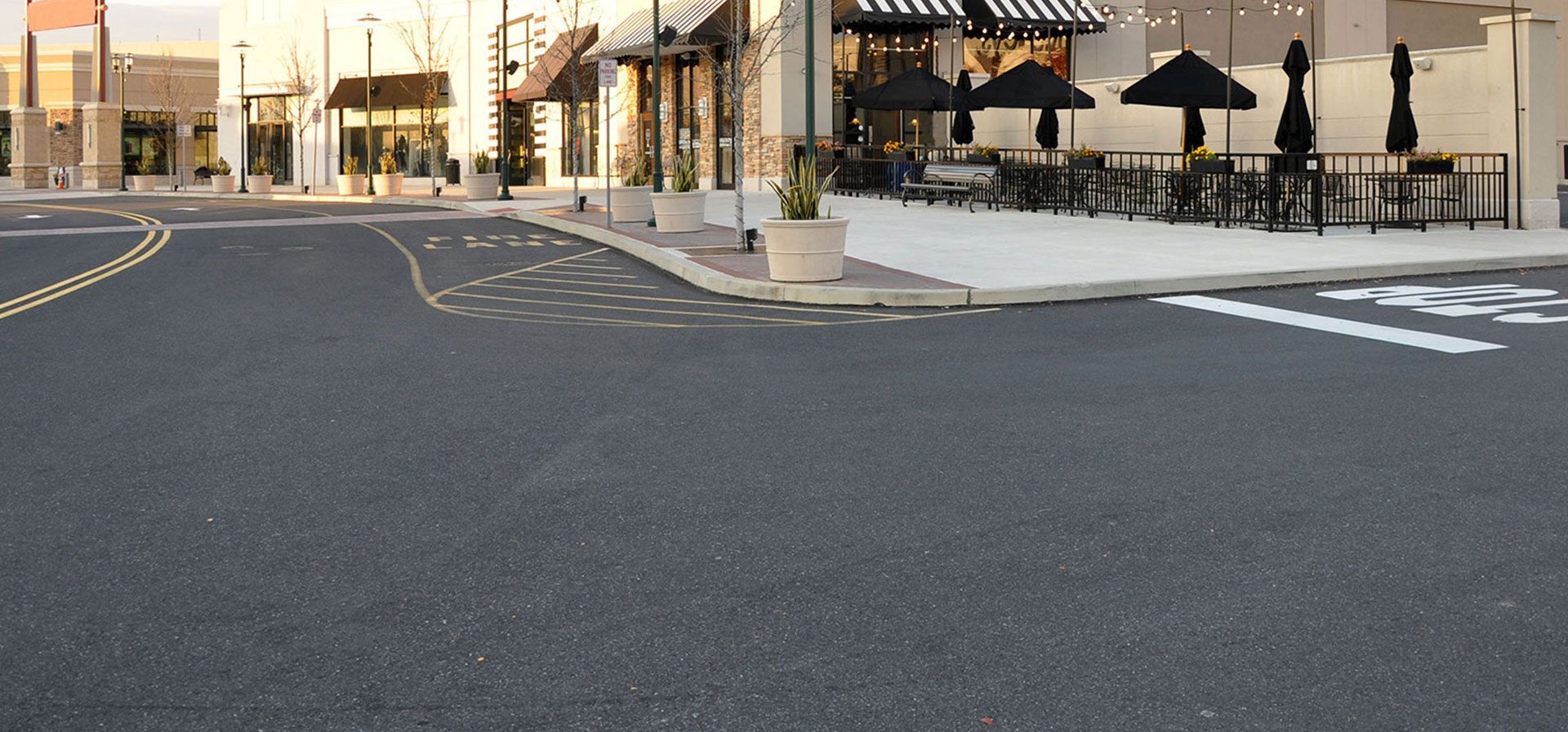 An asphalt parking lot in front of retail storefronts featuring an outdoor dining area with black tables and umbrellas.