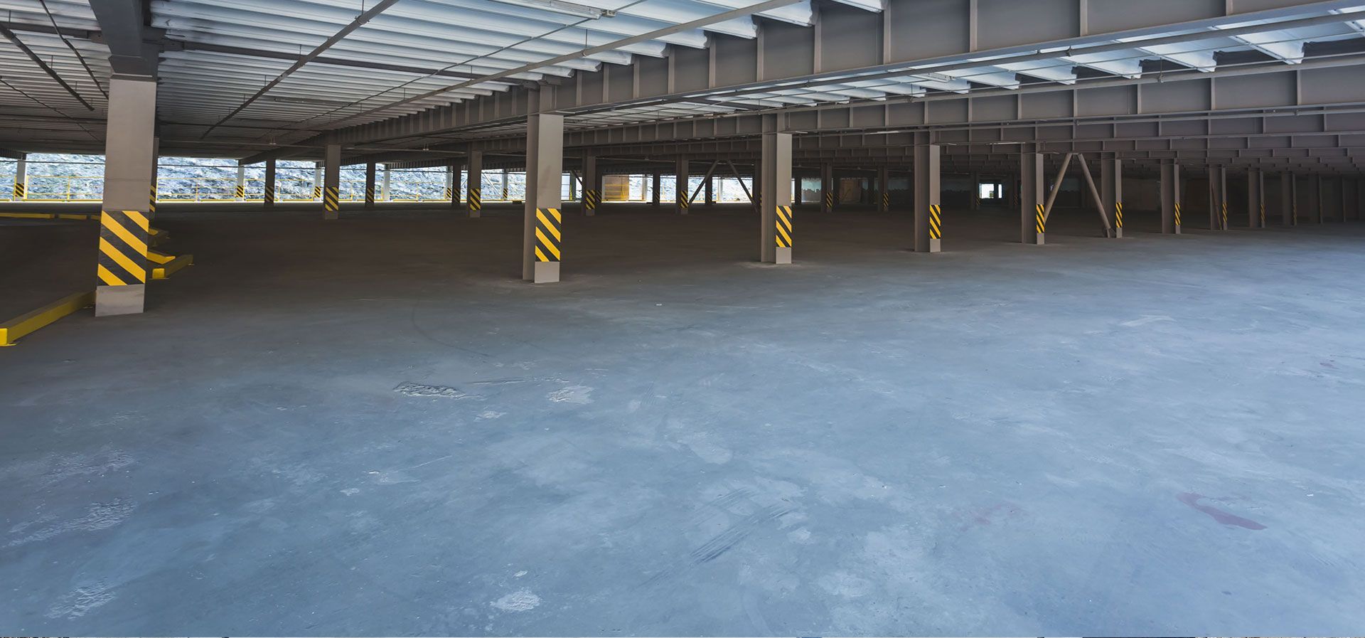 An empty indoor parking garage with grey concrete floors and support pillars with yellow and black hazard stripes.