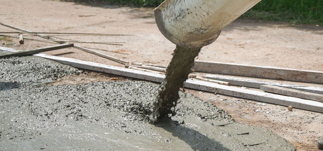 Wet concrete pours from a chute onto a construction site with wooden frame boards in the background.