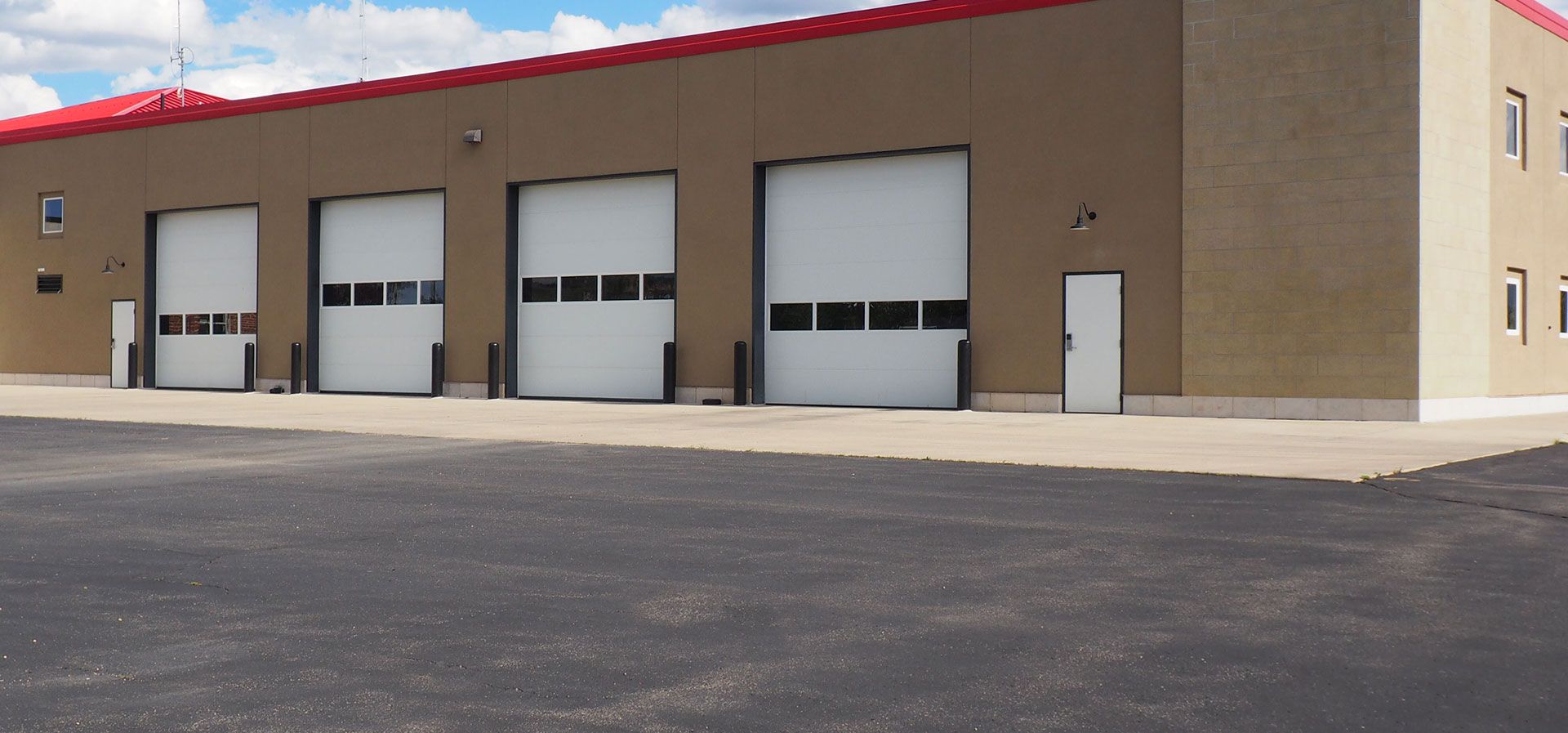 Tan industrial building with four large white garage bays and a side entry door under a red roof against a blue sky.