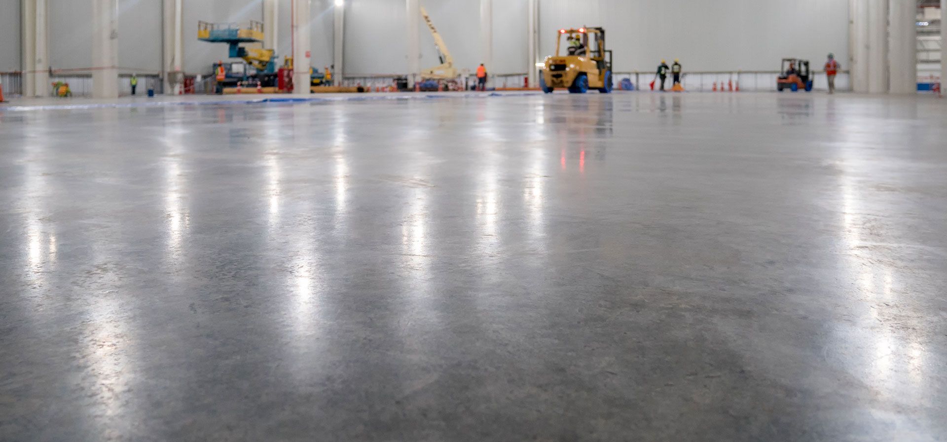 Shiny polished concrete floor in a large, bright industrial warehouse with construction equipment and workers in the back.