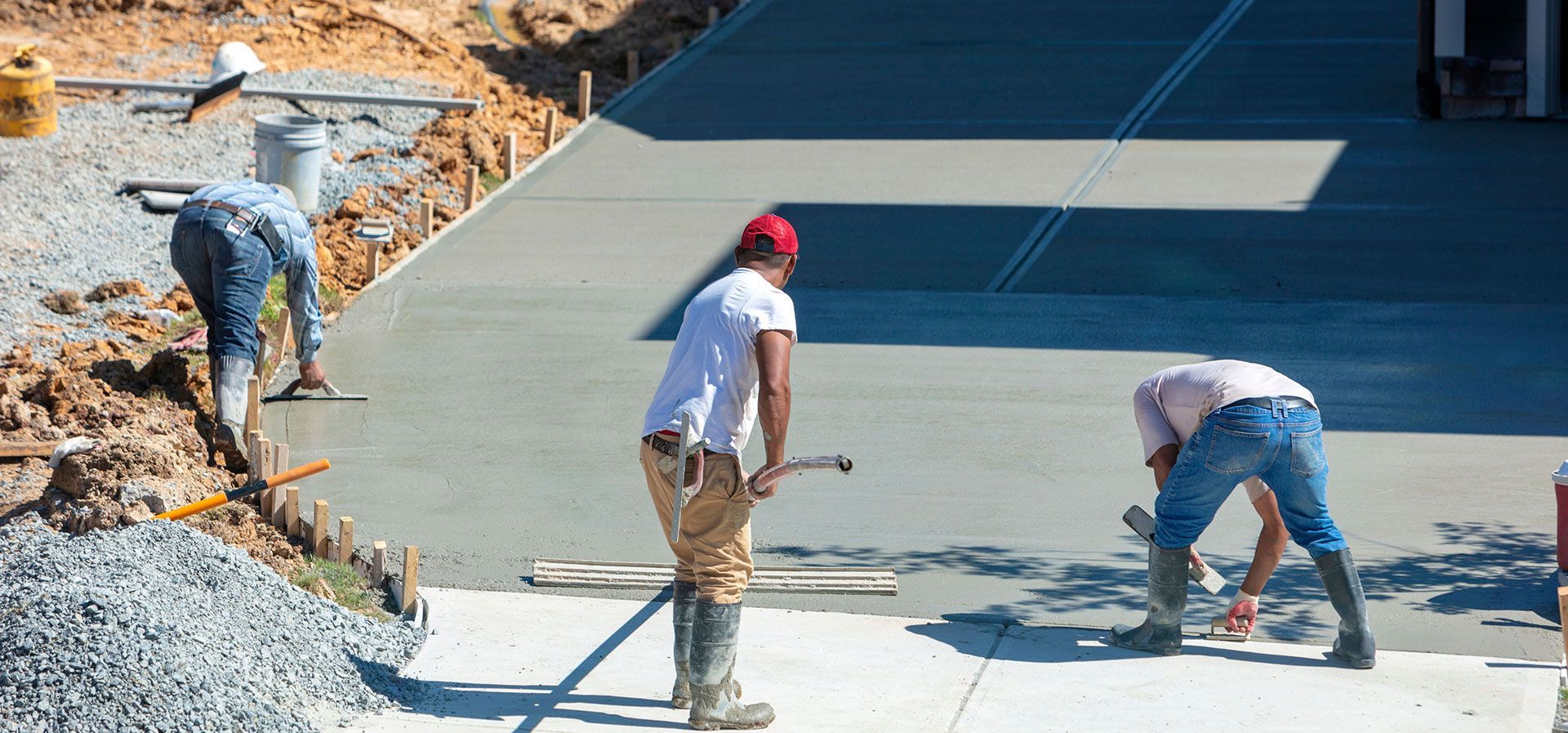 Three construction workers smooth and level fresh concrete on a driveway at an outdoor job site.