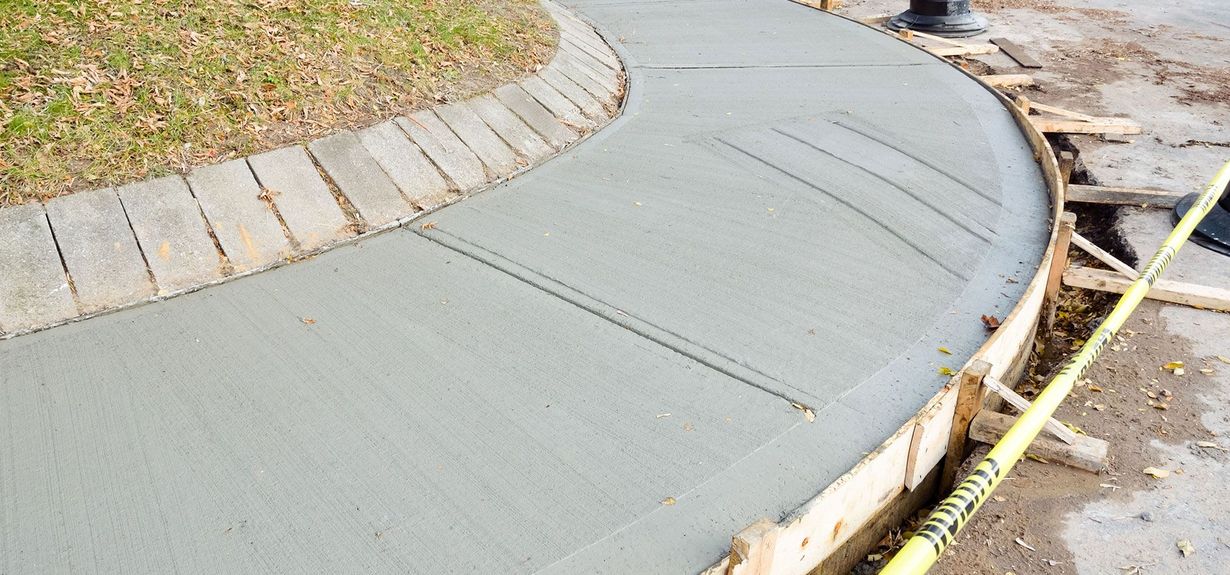 A freshly poured concrete sidewalk curves alongside a border of brick pavers and wooden construction forms.