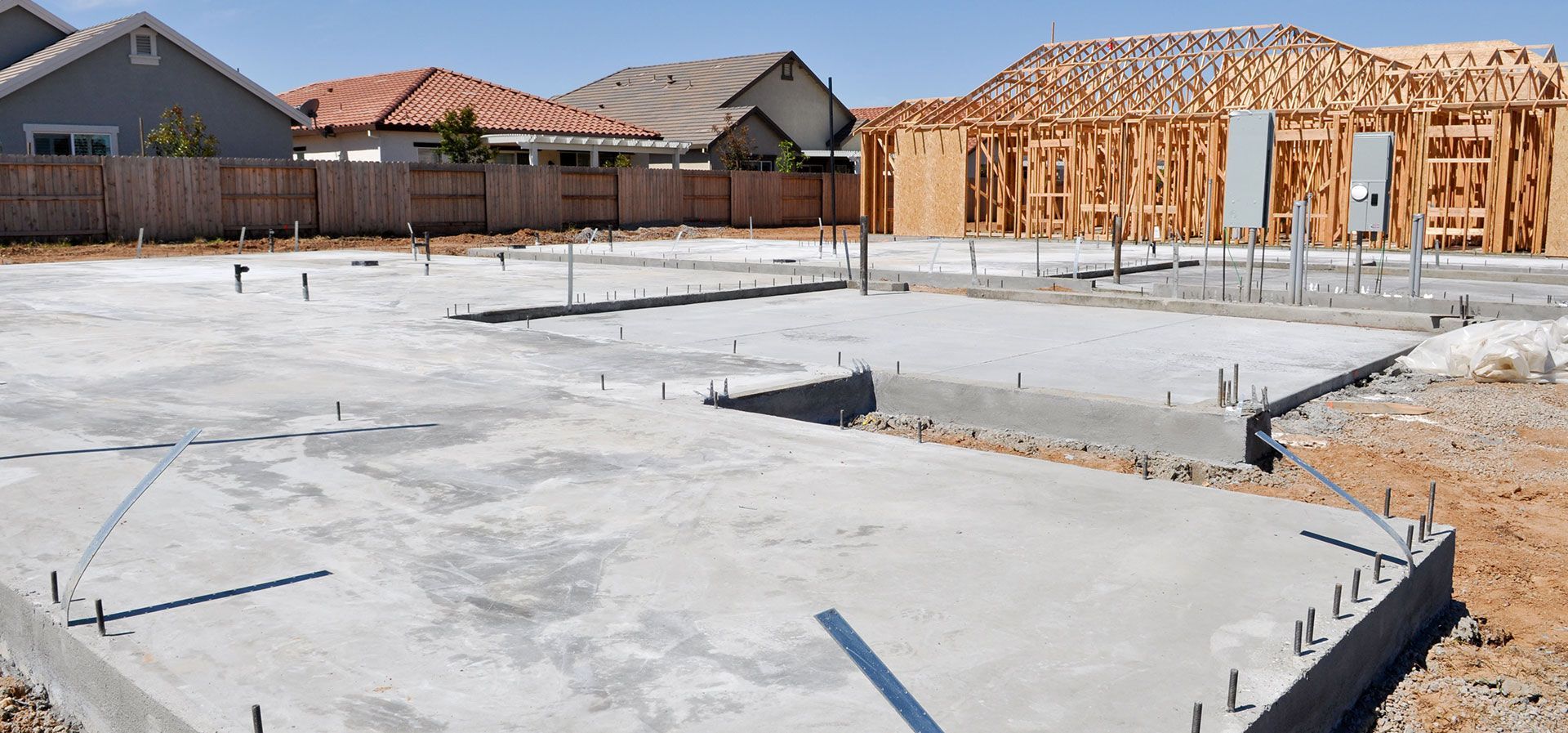 A concrete foundation slab at a residential construction site with wooden framing visible for a nearby house.