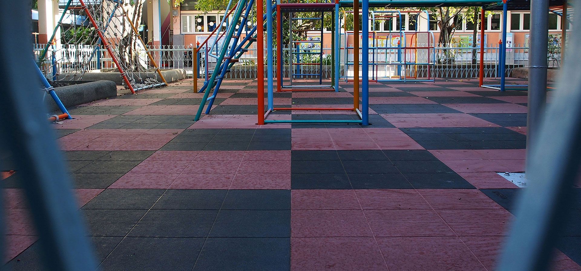 A playground with a black and red checkered rubber floor, viewed through the metal bars of a fence.