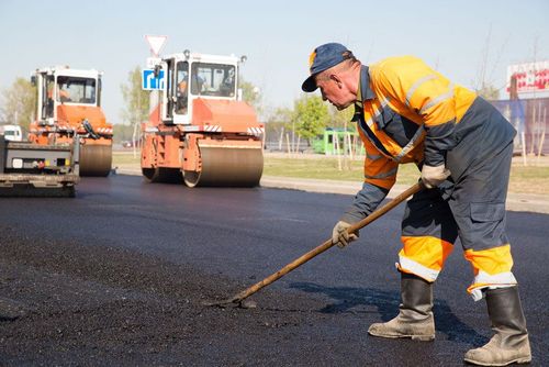 A construction worker in high-visibility gear spreads asphalt on a road while rollers operate in the background.