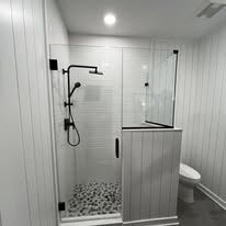 A modern bathroom with a glass-enclosed shower featuring a black rain shower head, pebbled floor, and white paneled walls.