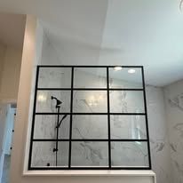 A glass shower partition with a black grid frame against a marble-tiled wall in a modern bathroom.