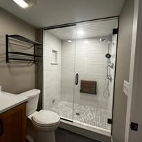 A modern bathroom featuring a glass-enclosed shower with white subway tile, a gray tiled floor, a toilet, and wall shelves.