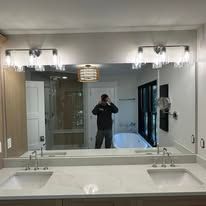A person takes a mirror selfie in a modern, bright bathroom with double sinks and two glass-shaded light fixtures.