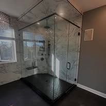 A modern walk-in shower with black-rimmed glass doors, marble-patterned walls, and a dark, textured tile floor.