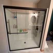 A walk-in shower with white walls, a glass door, dark metal trim, and built-in grab bars and storage shelves.