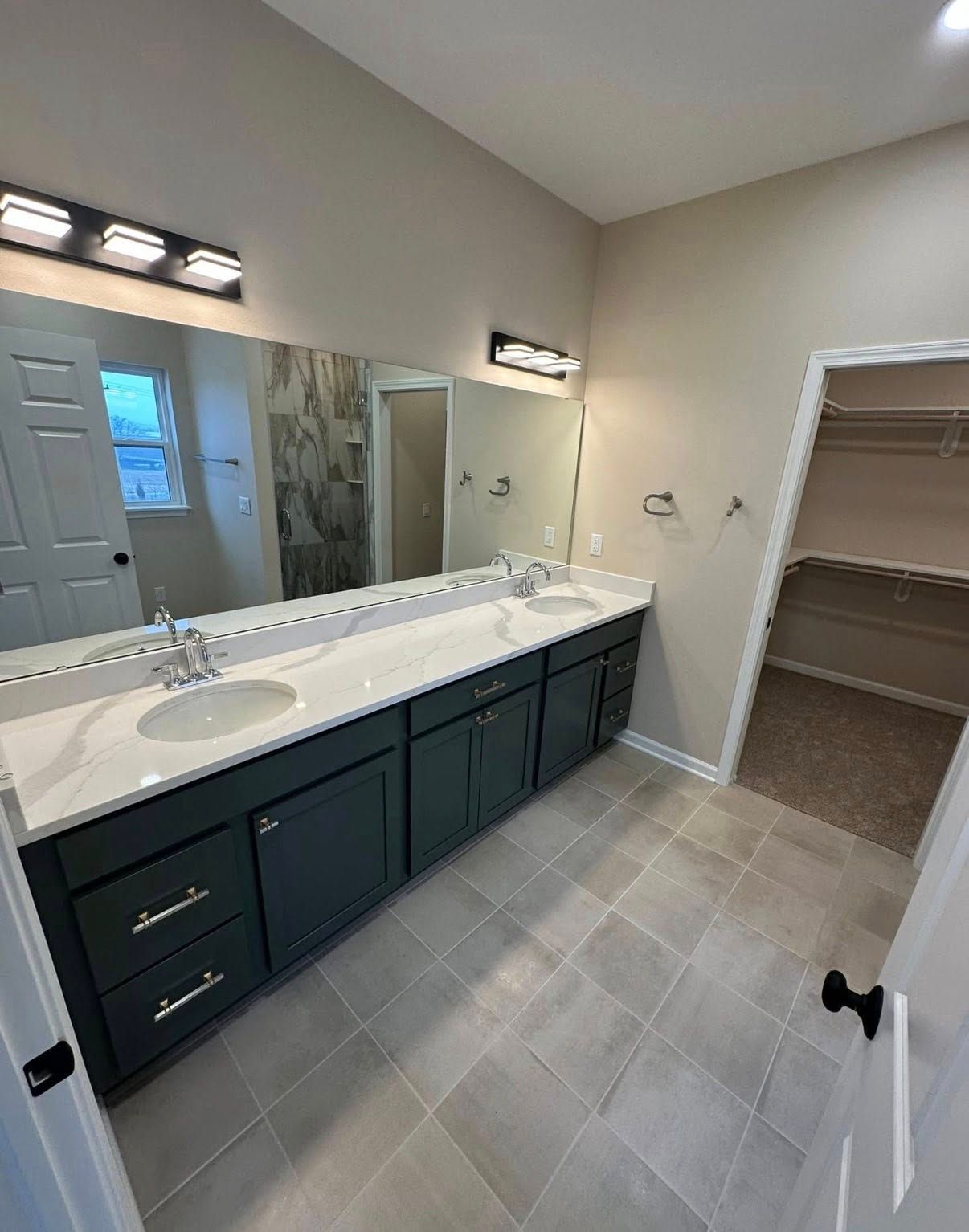 A bathroom with a double vanity featuring dark green cabinets, white countertops, two sinks, mirrors, and a walk-in closet.