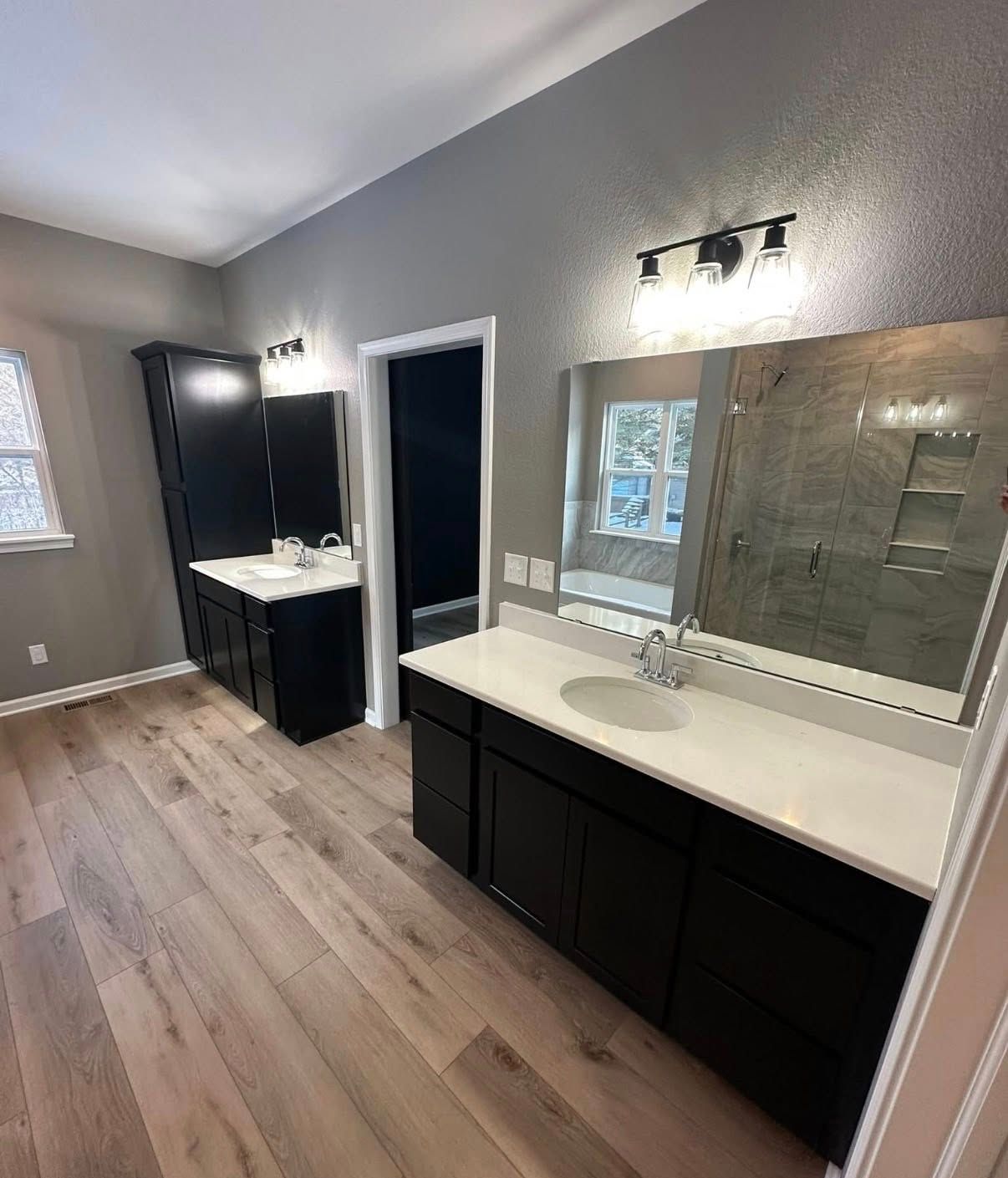 A modern bathroom features dark cabinets, white countertops, a large mirror, and wood-look flooring under grey walls.