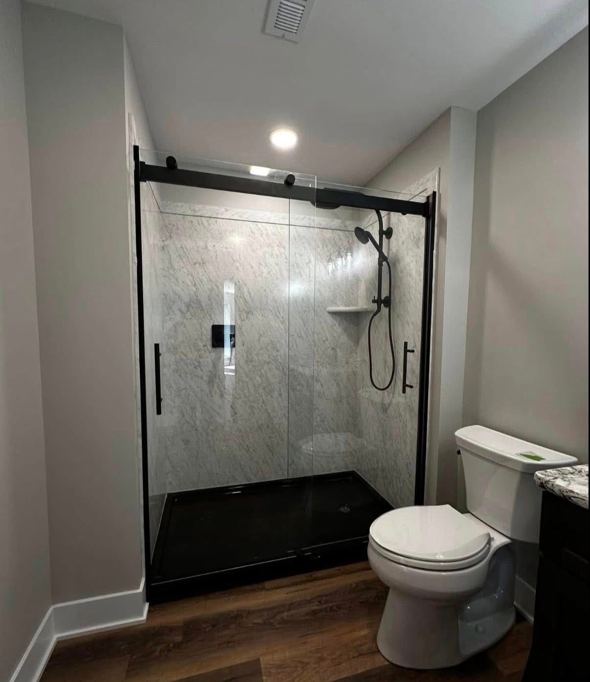 A bathroom with a black-framed sliding glass shower, marble-patterned tile, a white toilet, and dark wood-look flooring.