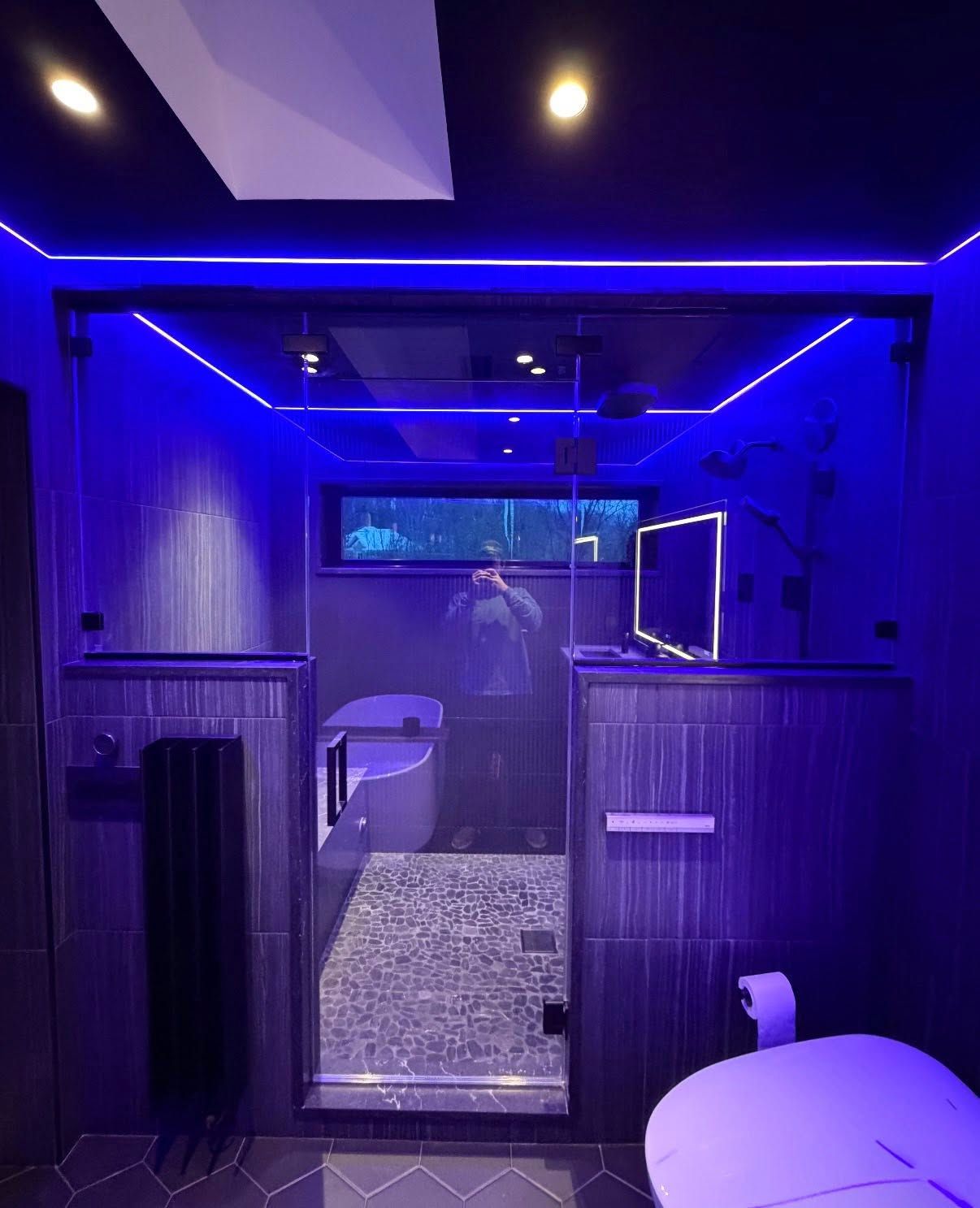 Modern bathroom with blue ambient lighting, a walk-in glass shower, a bathtub, and dark textured wall tiles.