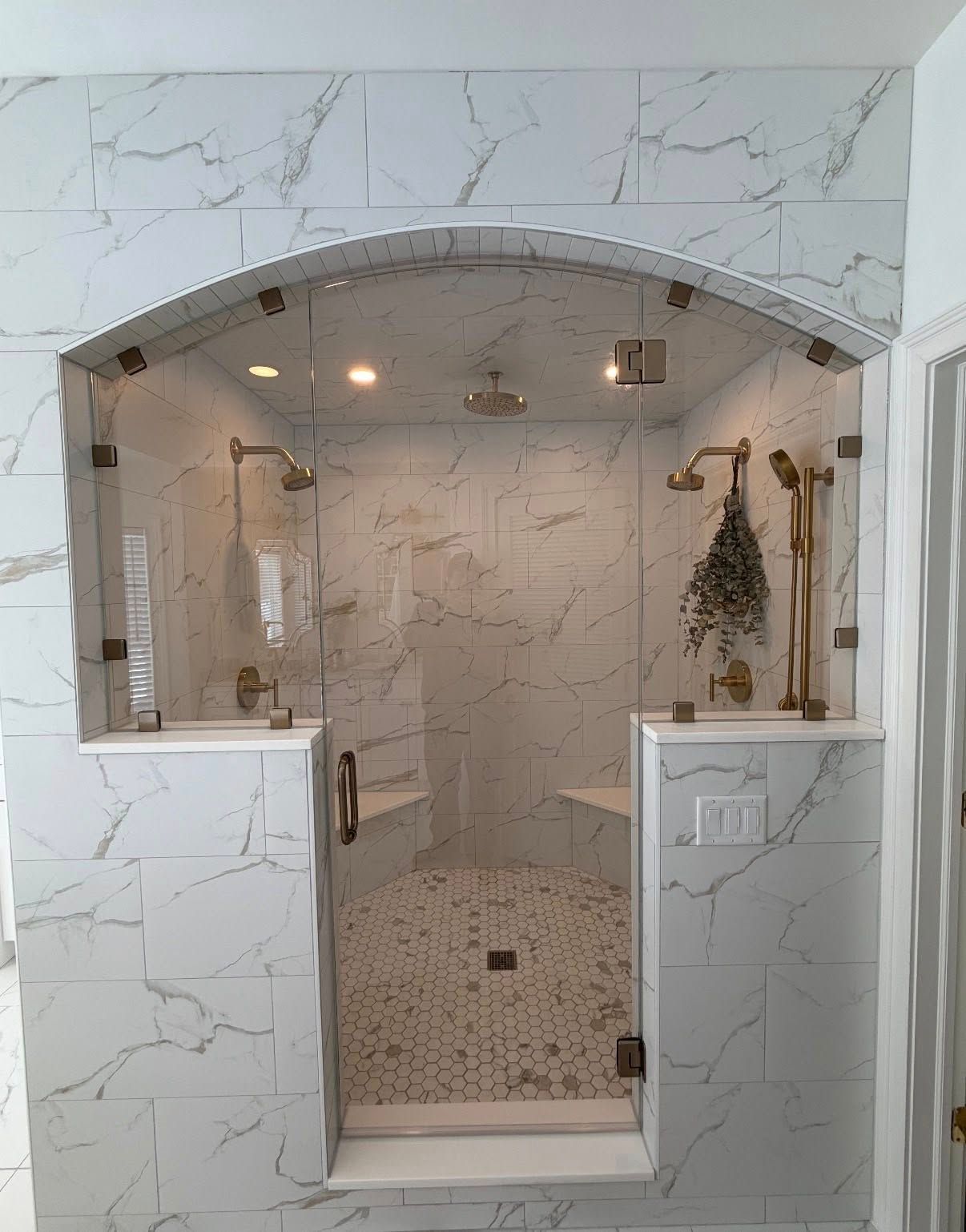 A walk-in shower with white marble-patterned tile, glass door and walls, two shower heads, and a pebble-tiled floor.