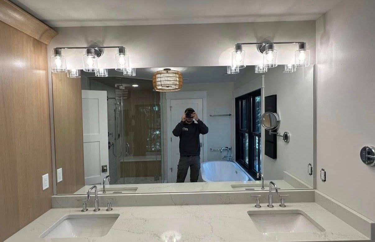 A double-sink bathroom vanity with two light fixtures above a large mirror reflecting a person taking a photo.