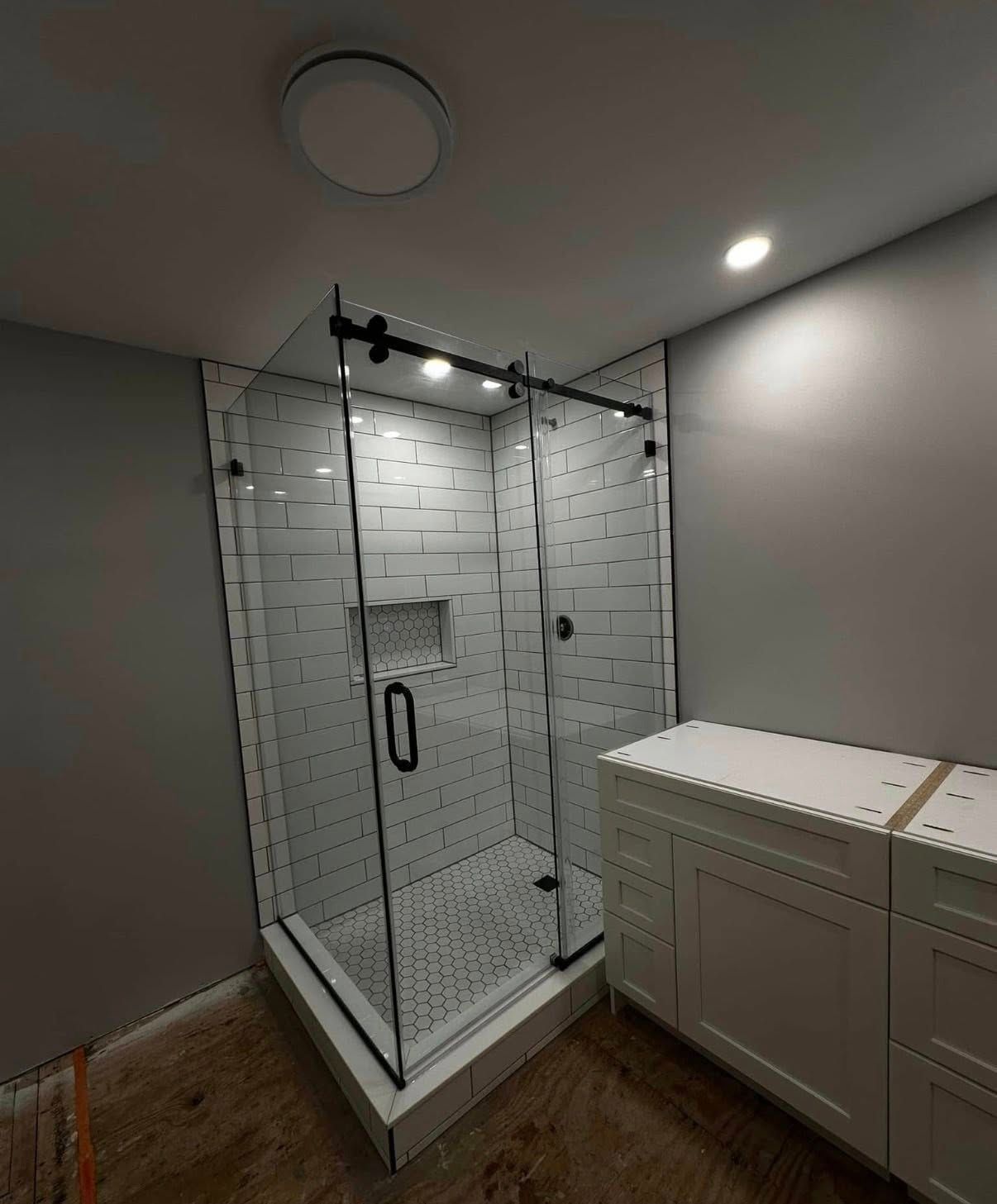 A modern, white tiled corner shower with a glass sliding door and a vanity next to it in an unfinished room.