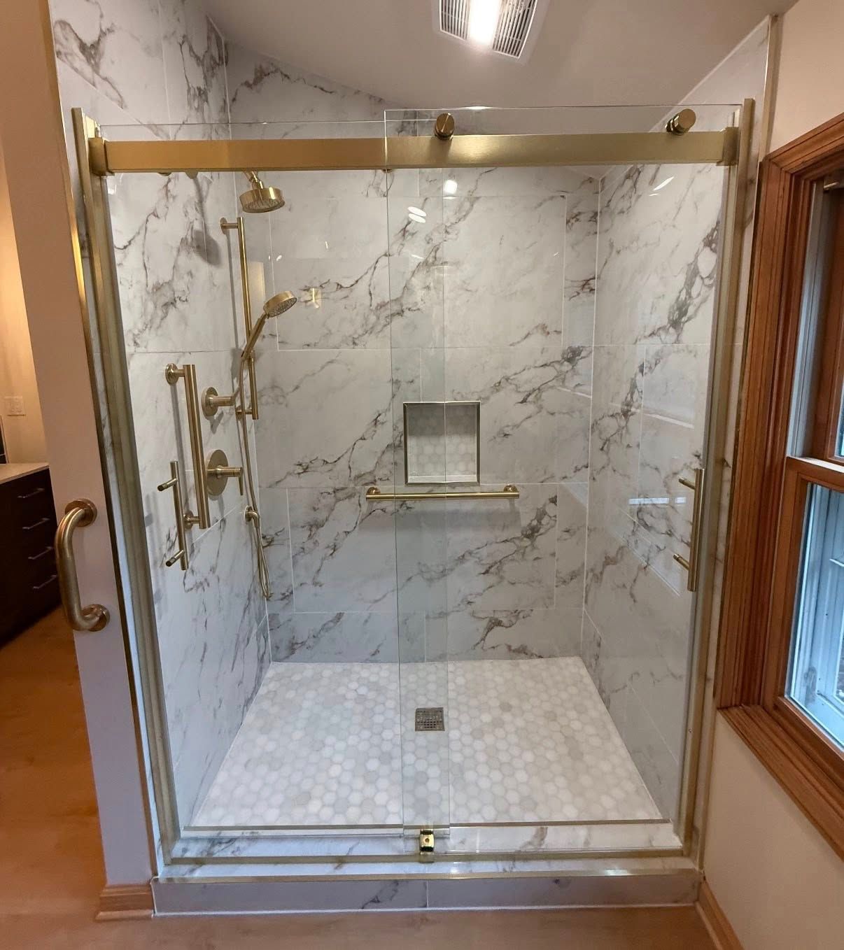 A walk-in shower with white marble-patterned tile, gold fixtures, a glass sliding door, and a pebble-tiled floor.