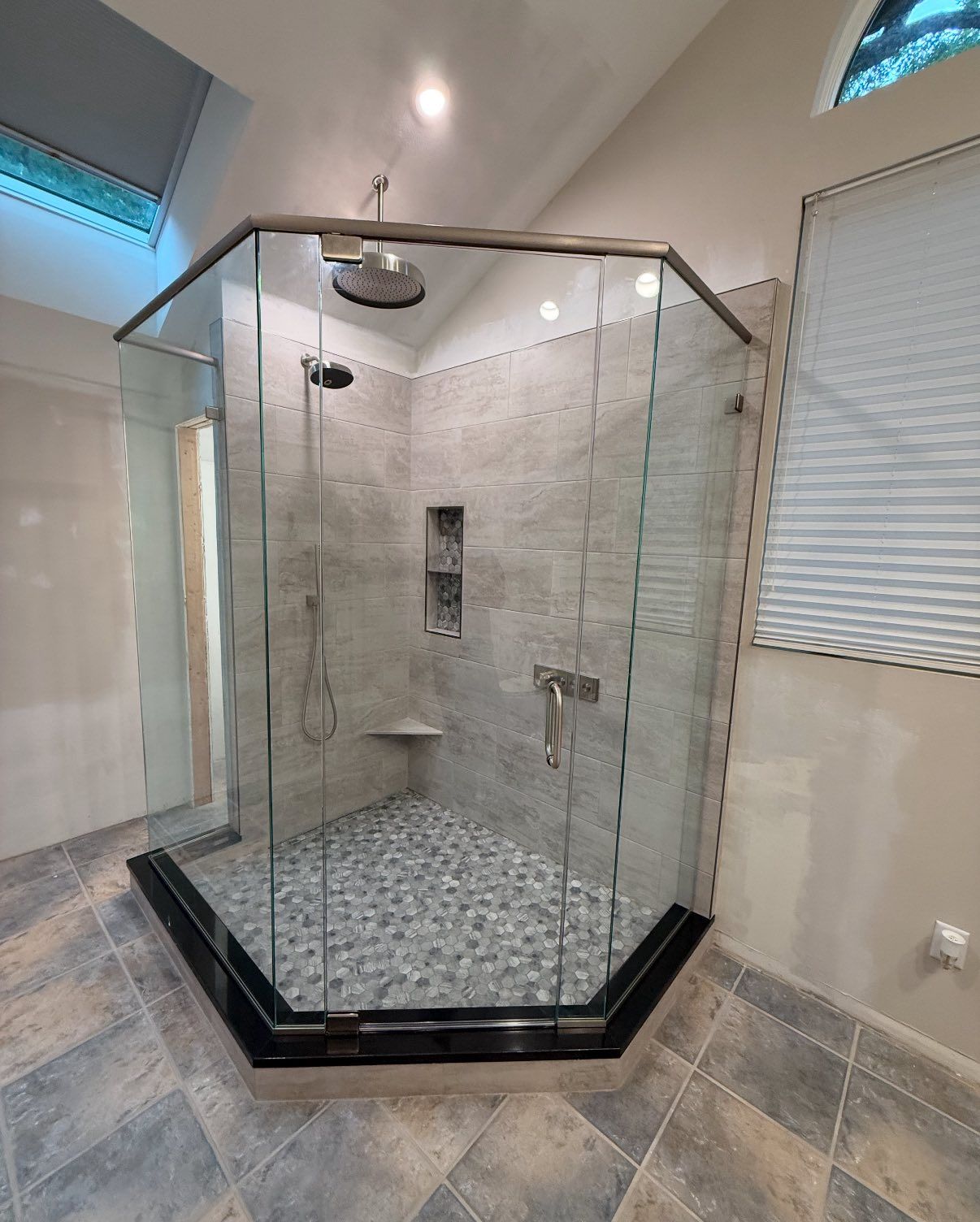 A modern corner shower with a glass enclosure, pebble-stone floor, gray tile walls, and a black base in a bright bathroom.