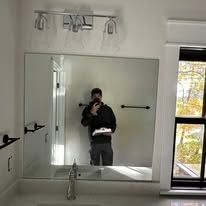A person in a black hoodie takes a selfie in a bathroom mirror beneath a three-light vanity fixture.