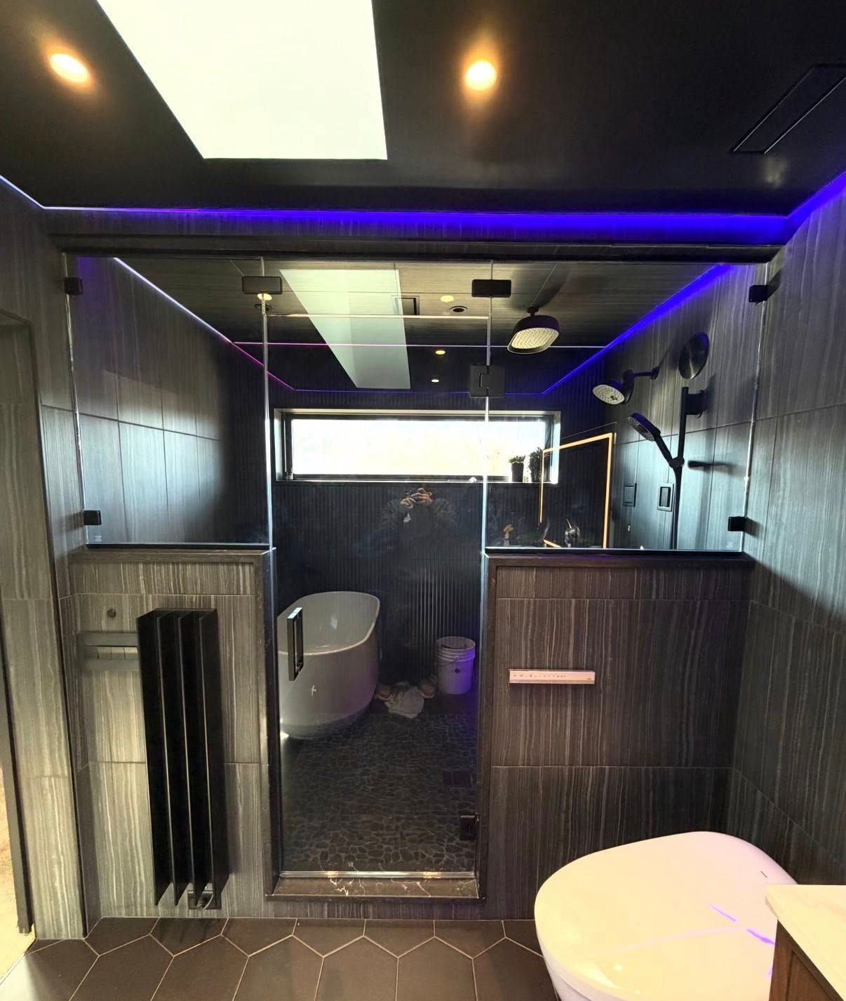 A modern bathroom with dark, textured tile walls, purple LED accent lighting, a glass-enclosed shower, and a soaking tub.