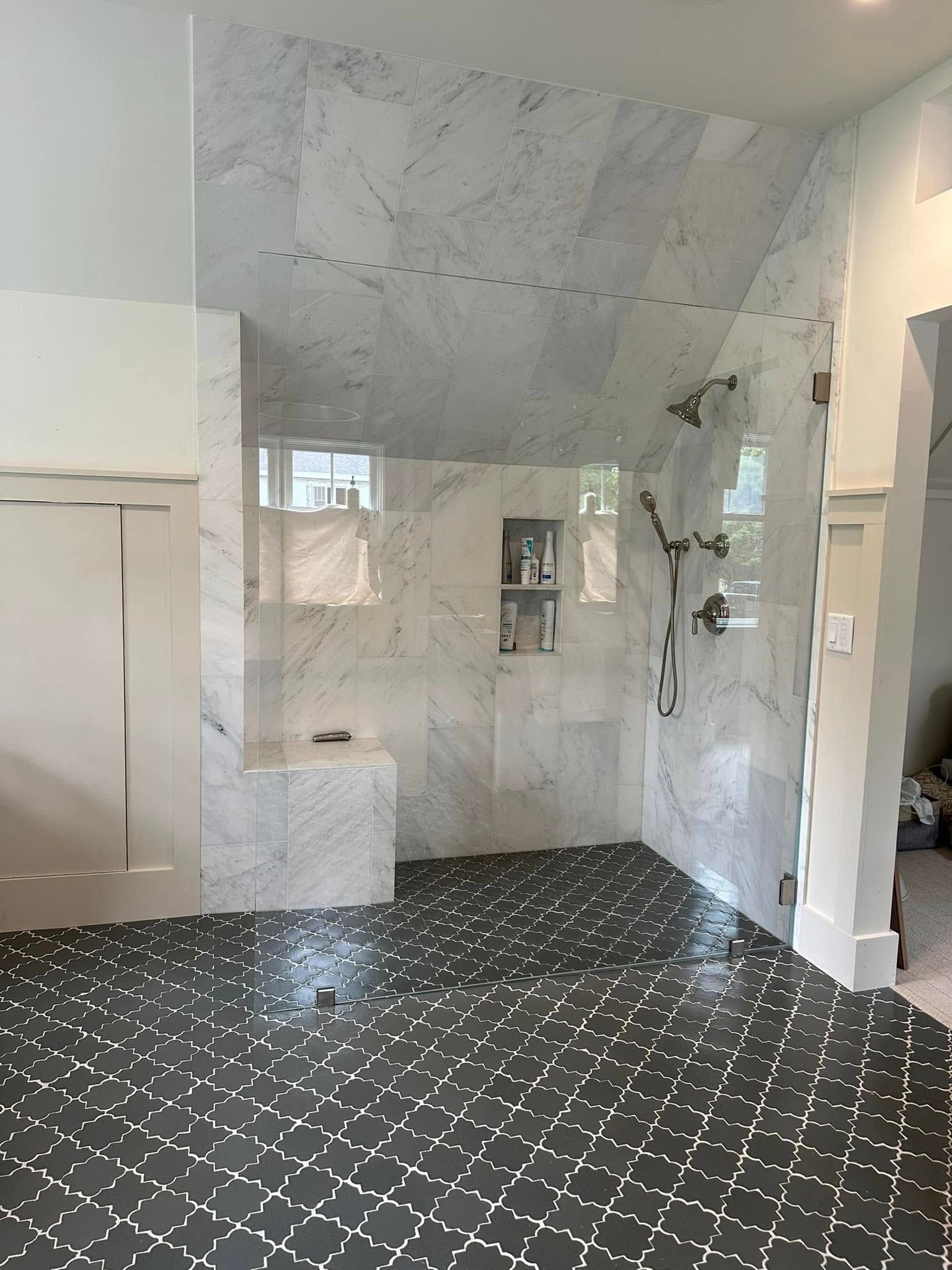 A shower with white marble walls and a built-in bench, featuring dark patterned floor tiles and a glass door.