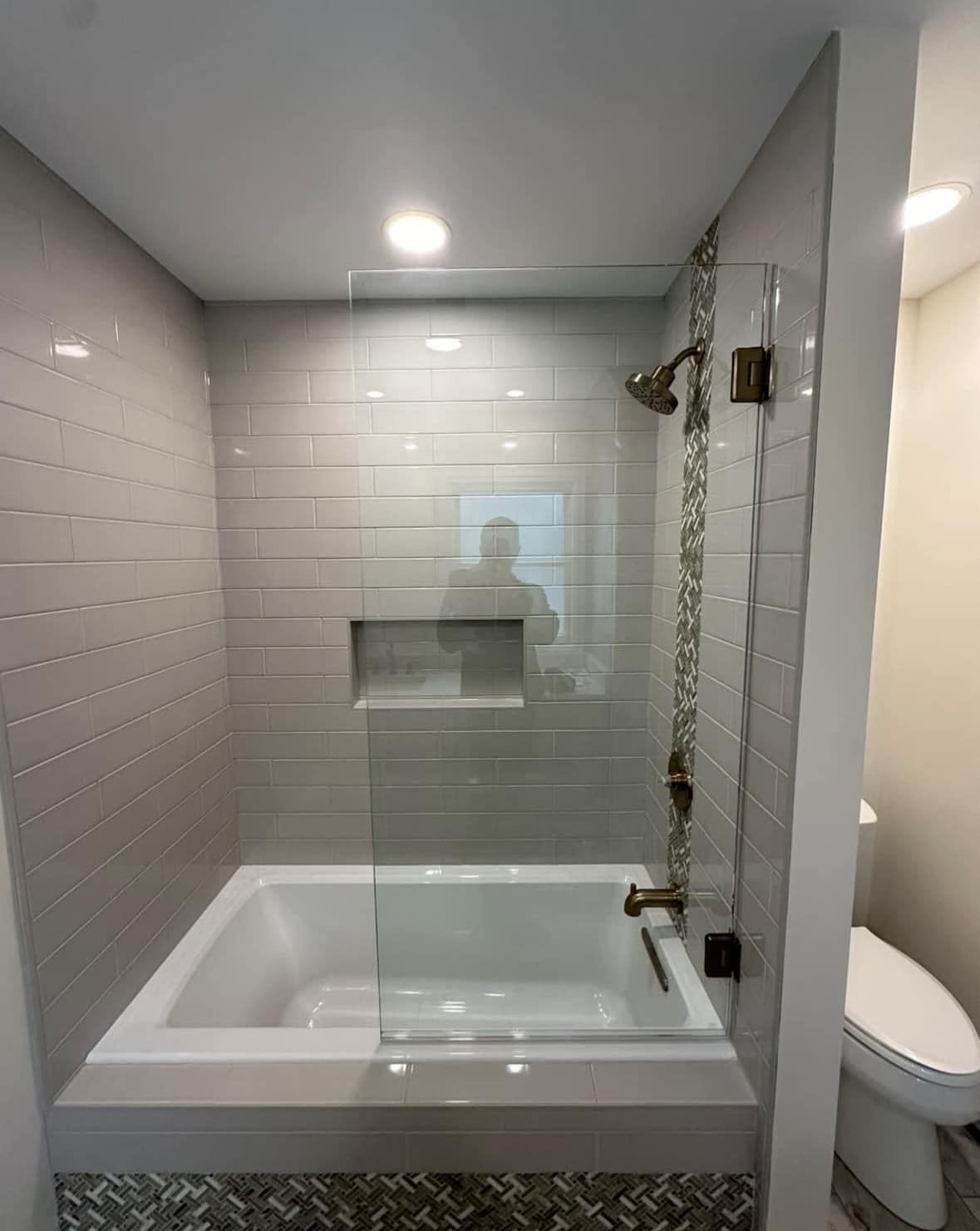 A modern bathroom with a white soaking tub, light gray subway tile walls, a glass splash panel, and a niche for toiletries.