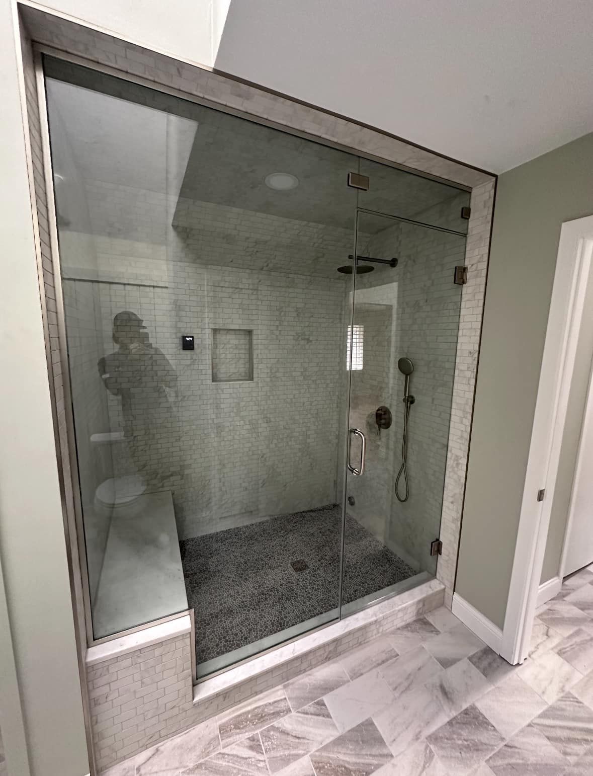 A modern walk-in glass shower featuring a stone bench, pebble-style flooring, and light marble wall tiles.